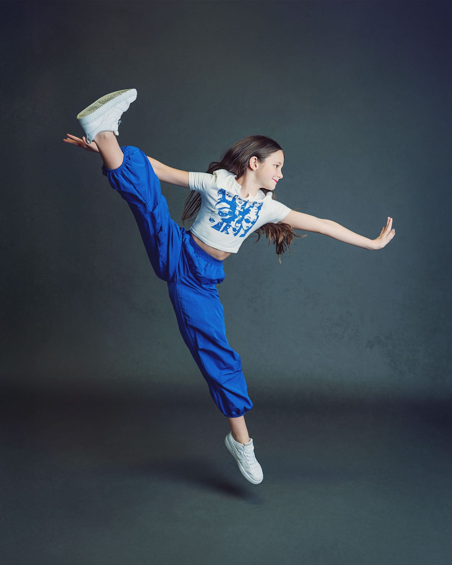 A dancer in a bright blue shirt performs a graceful arabesque pose against a dark studio backdrop.