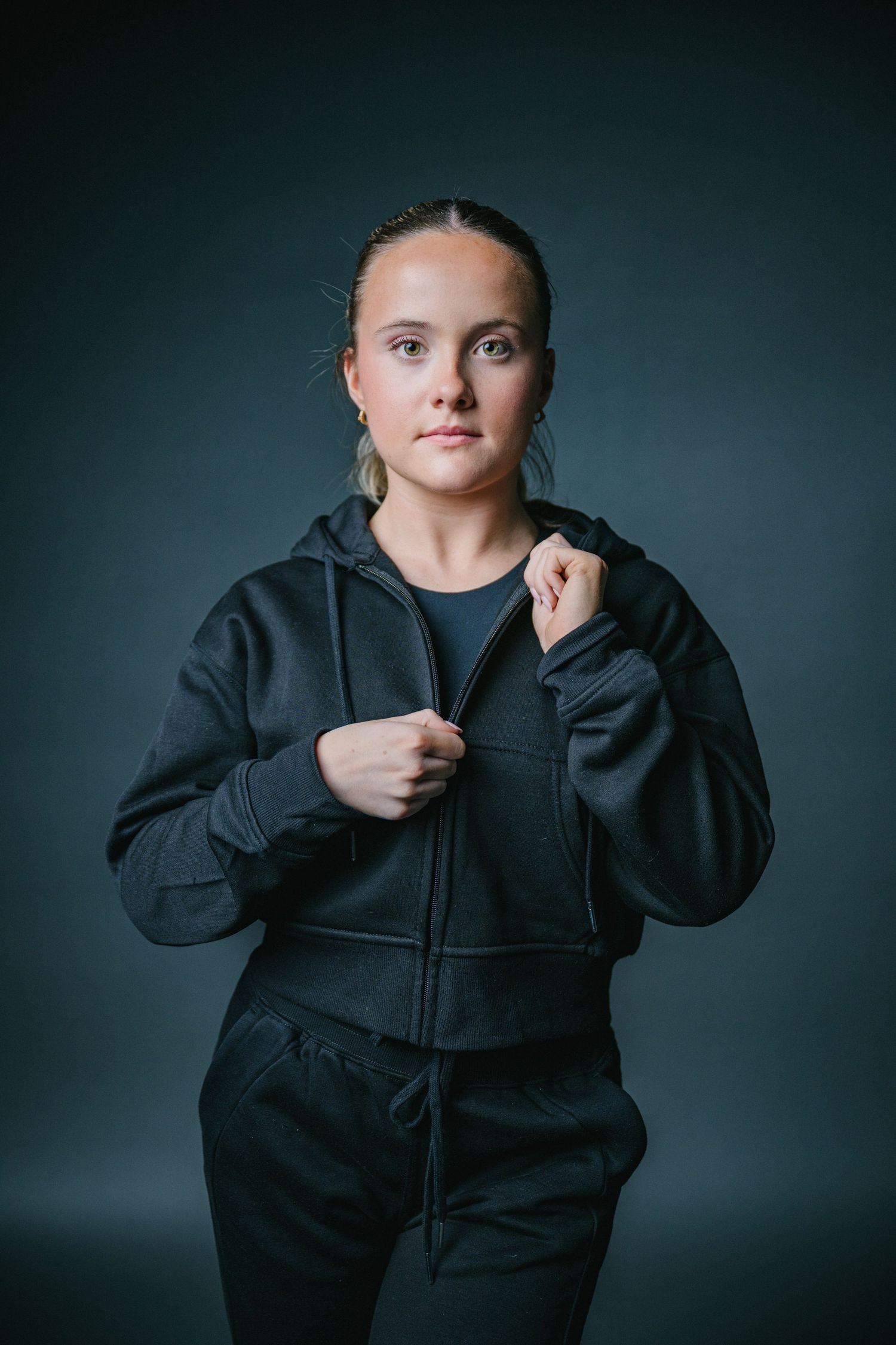 Figure in black hoodie shown in four sequential photos demonstrating how to zip up the garment against dark background.