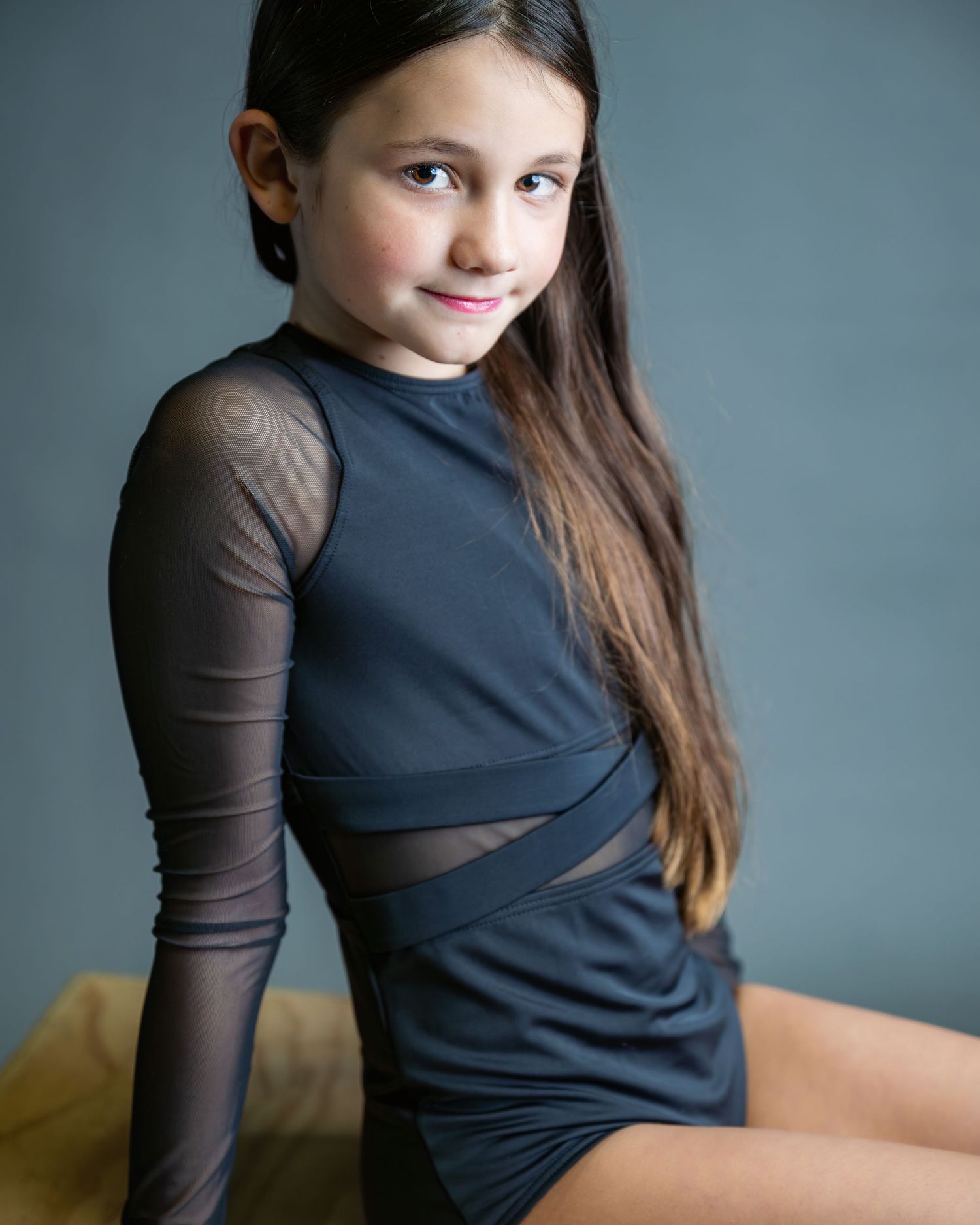 Young dancer wearing a black dance costume with sheer sleeves poses against a gray studio backdrop.