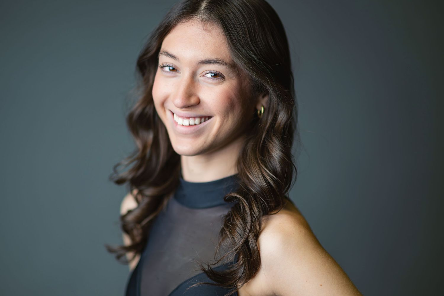 Professional studio headshot portrait with dark background showing warm smile and dark wavy hair.