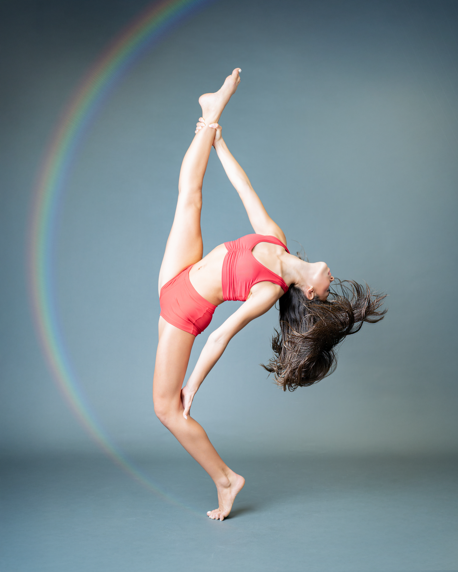 A dancer in a red leotard performs a graceful backbend pose with a rainbow arc visible in the background.