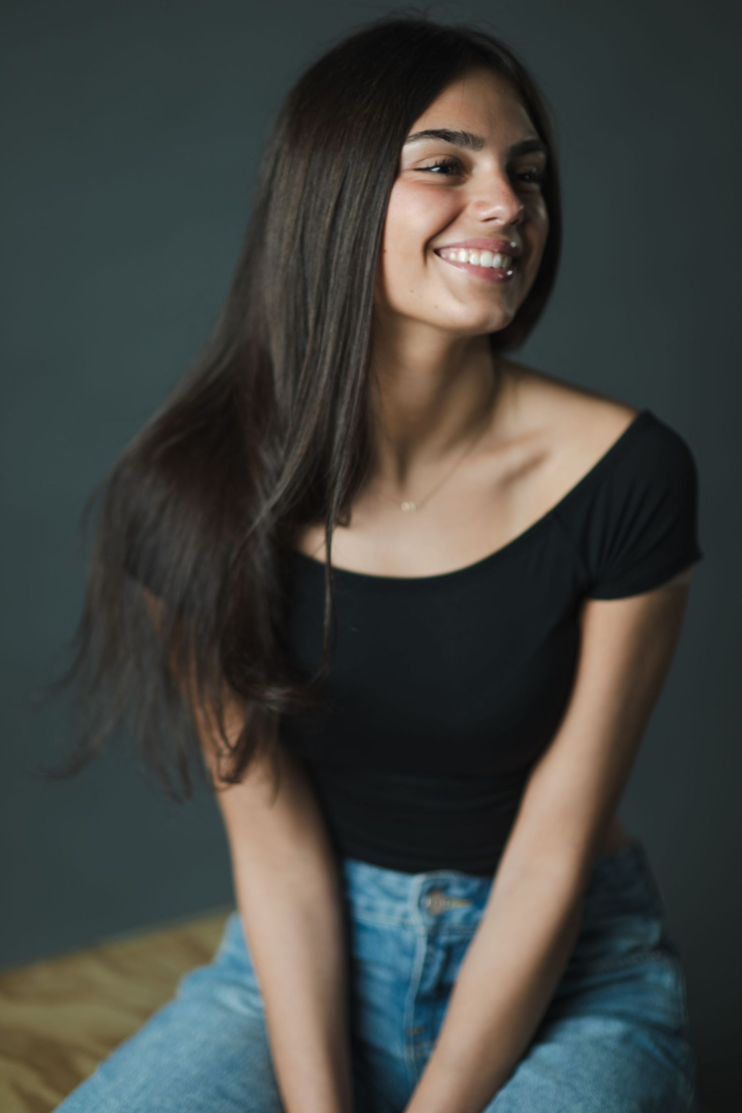 A joyful portrait against a dark green background wearing a black top and blue jeans with long flowing dark hair.