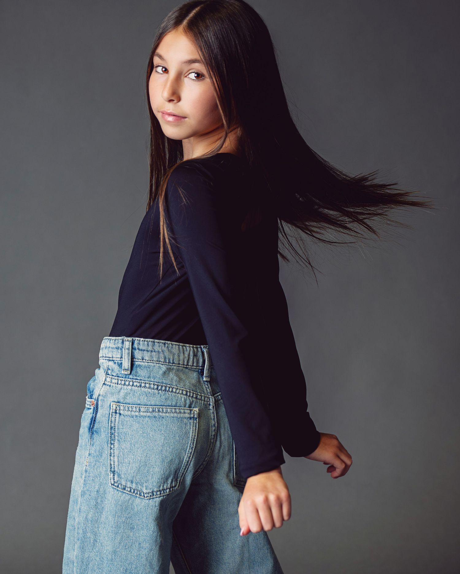 Fashion portrait series showing movement of long dark hair against grey background while wearing black top and light jeans.