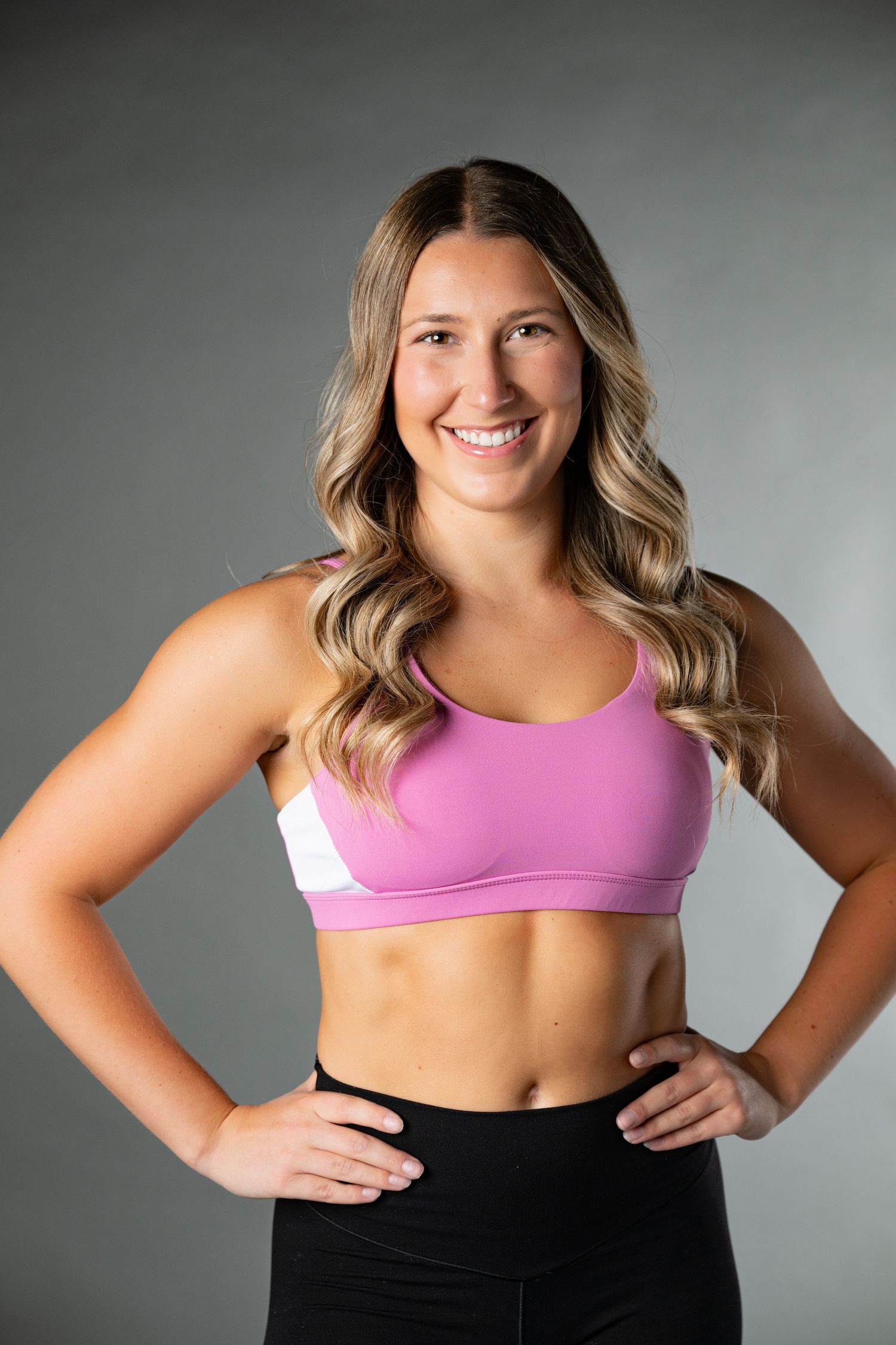 Athletic woman in pink sports bra and black leggings poses confidently against gray background in fitness photoshoot series.