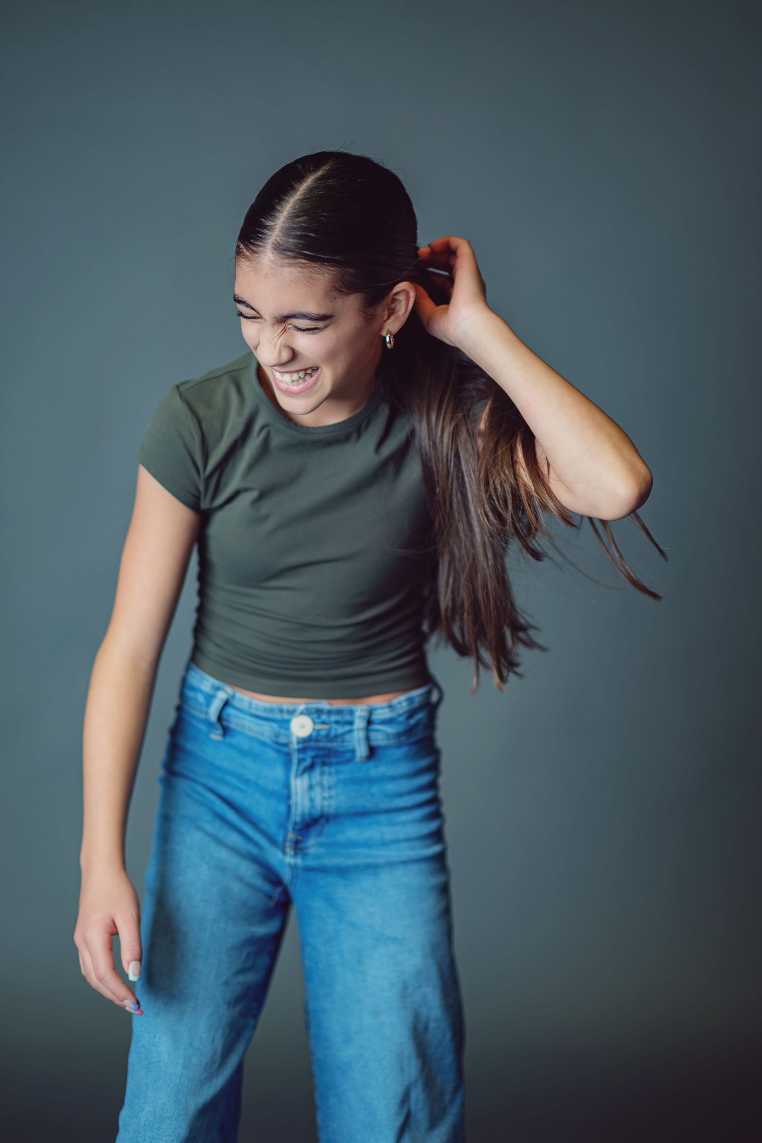 A woman in a dark green t-shirt and blue jeans moves playfully against a gray studio backdrop.