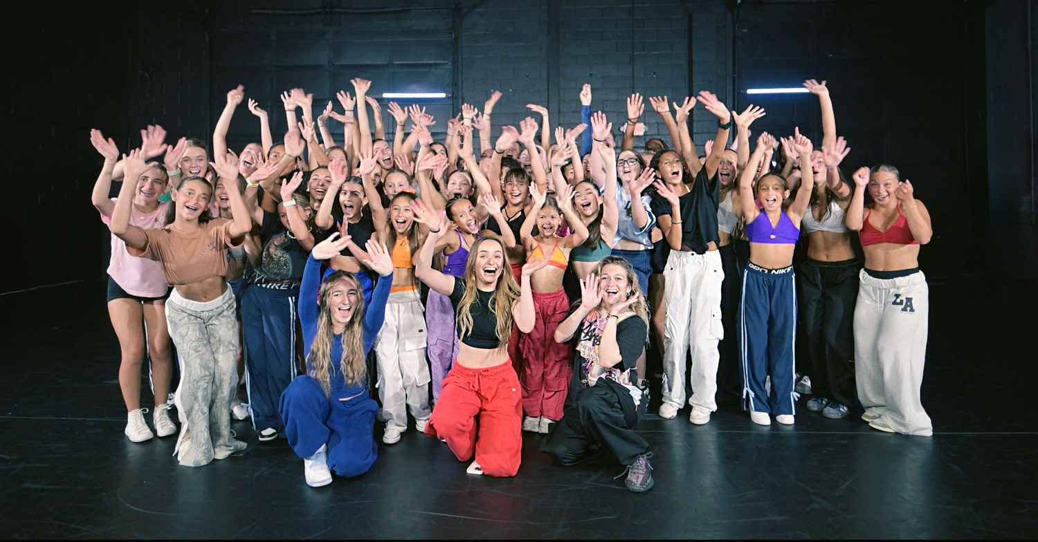 A large group of performers on stage raise their arms in celebration during a theatrical performance finale.