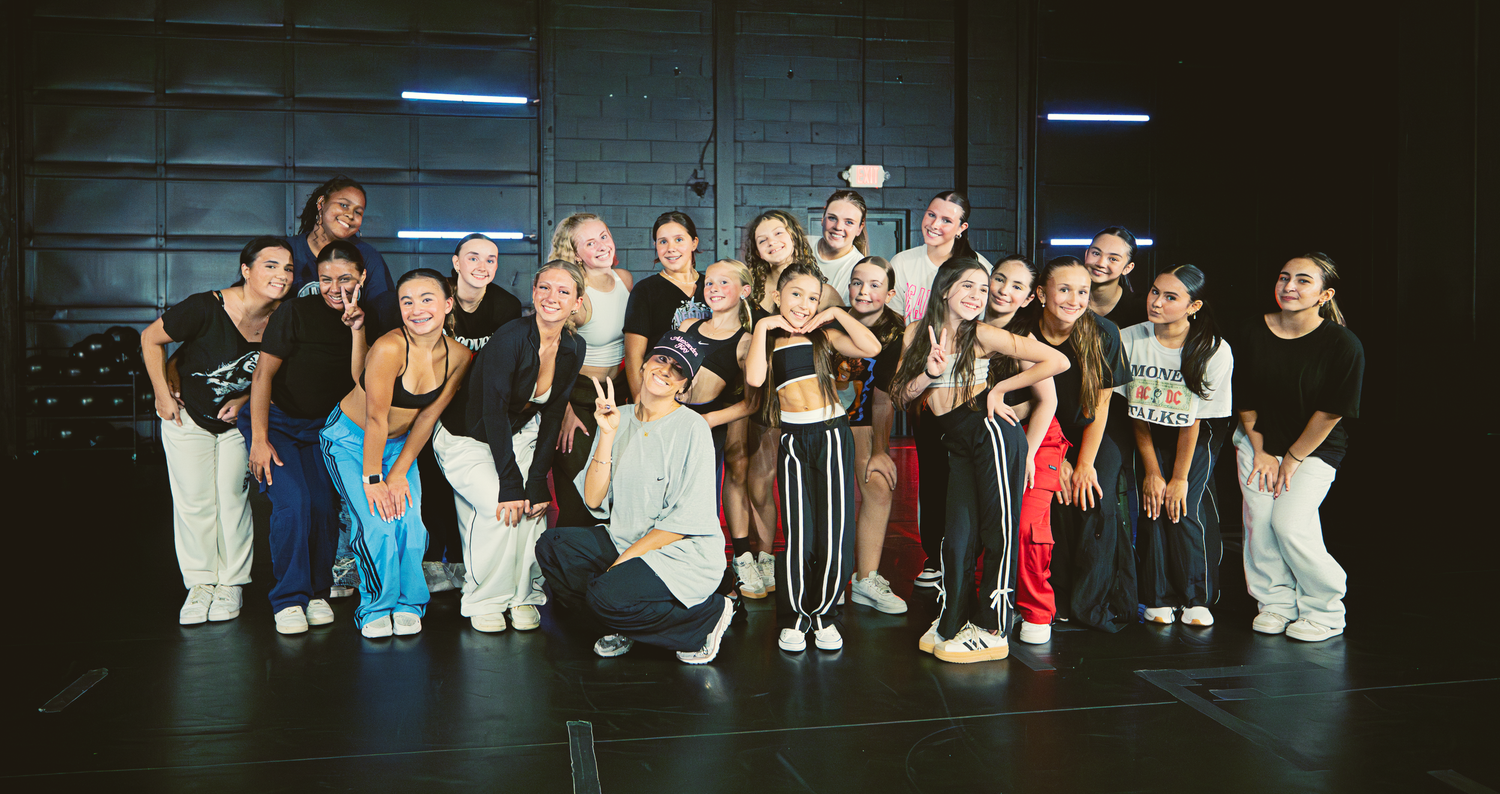 A large group of dancers poses together on a dark stage wearing casual dance attire and athletic wear.