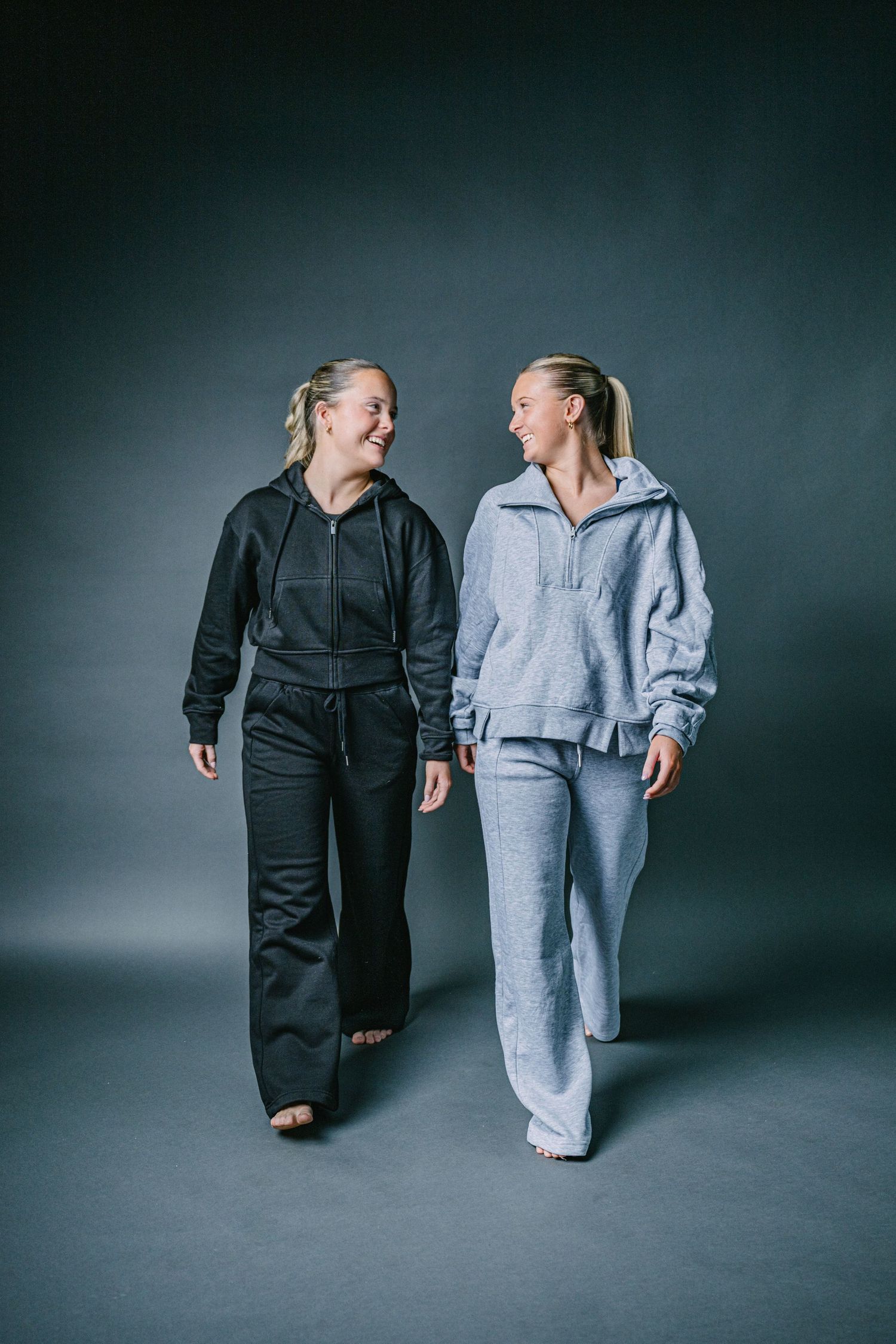 Two people wearing matching loungewear sets in black and gray walk together against a dark studio backdrop.