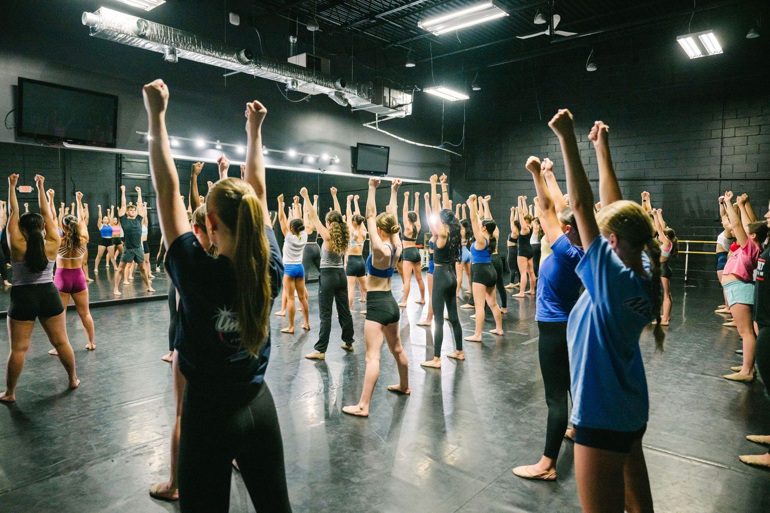 Large group of dancers practicing choreography in a spacious dark studio with fluorescent lighting.