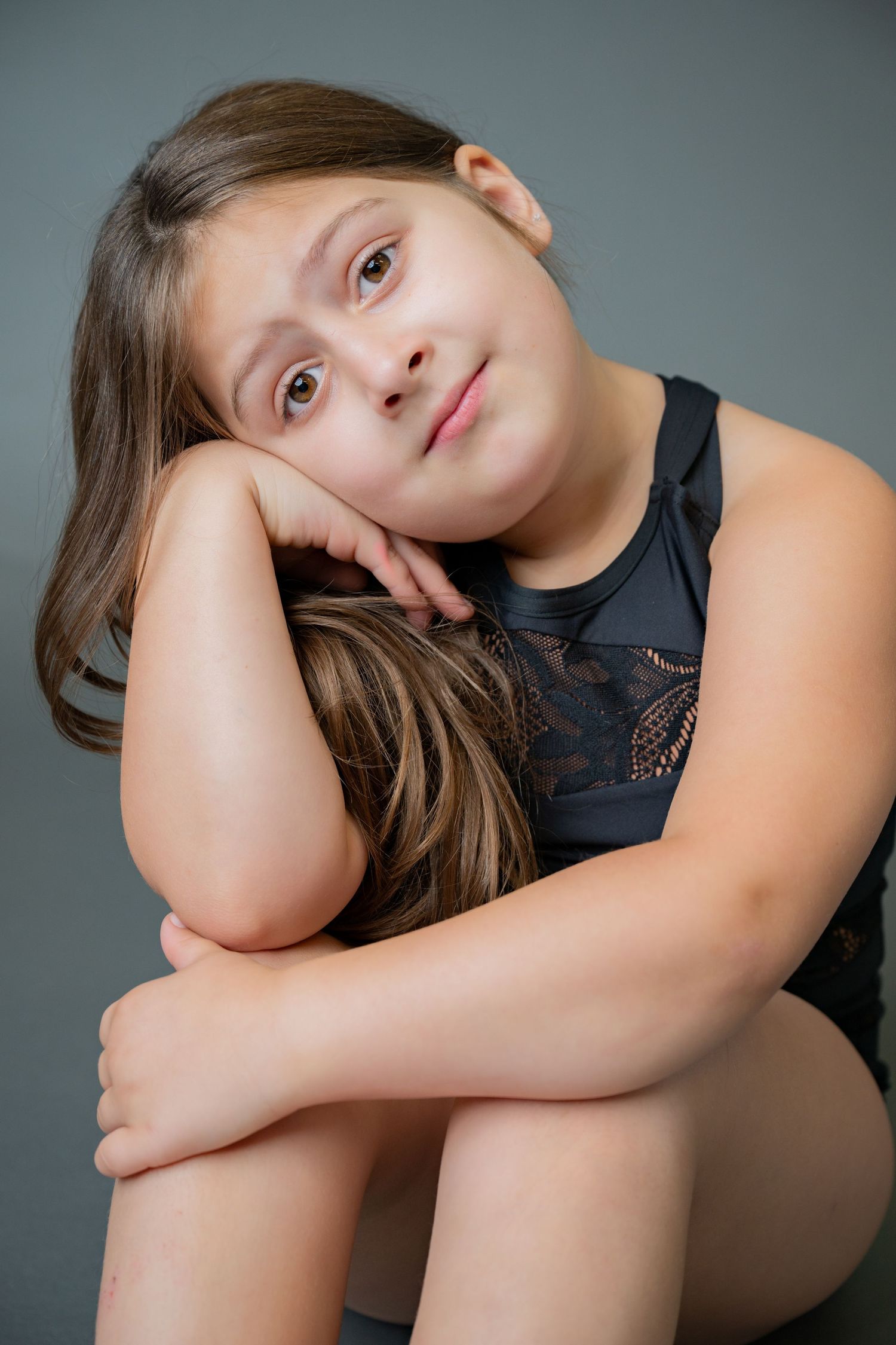 A child in a black top rests their head on their hand while sitting in a thoughtful pose against a gray background.
