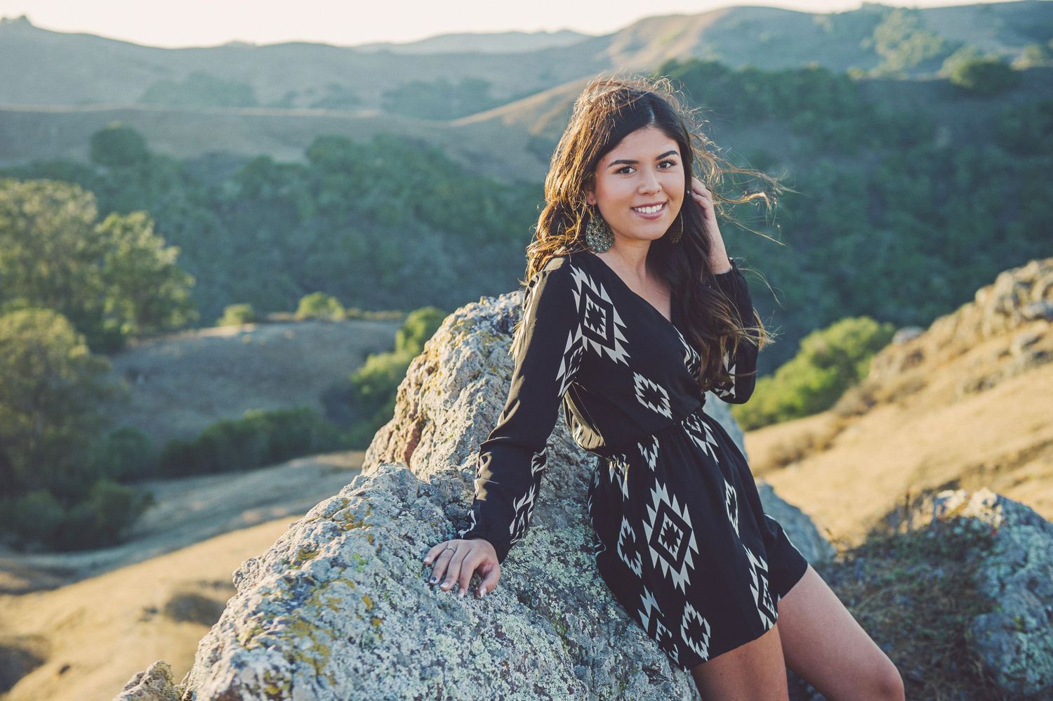A person in a patterned sweater sits on a large boulder overlooking rolling green hills on a sunny afternoon.