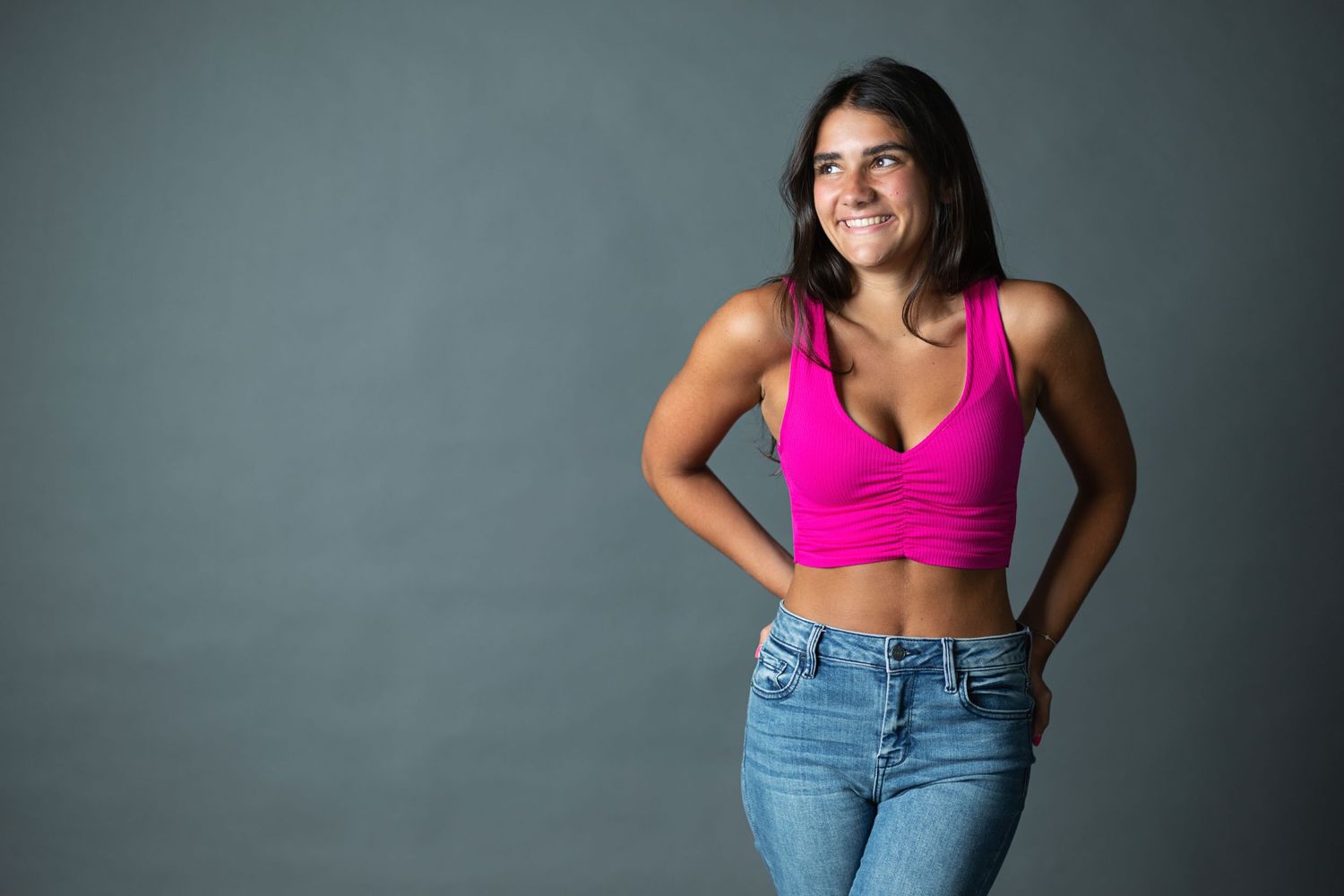 Person in blue jeans and pink sports bra poses against gray wall in six different poses showing body confidence.