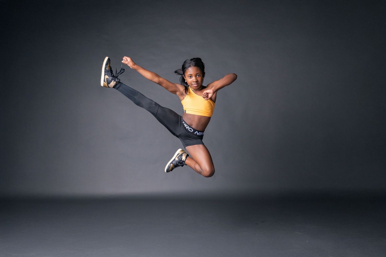 Athletic dancer in yellow top and black shorts performs dynamic split jump against dark studio background.