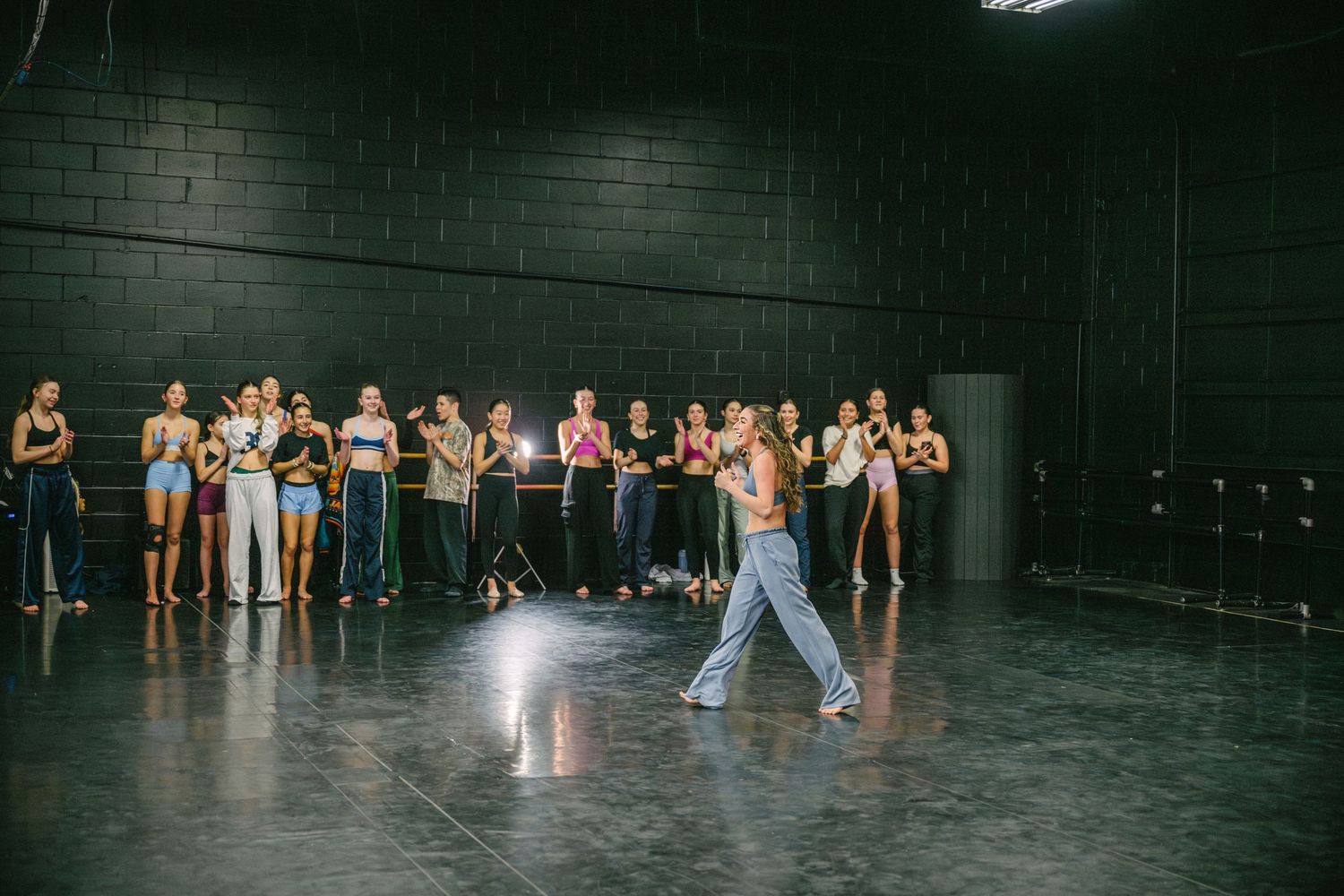 Dancers stretch and warm up together in a spacious dark studio with industrial lighting.
