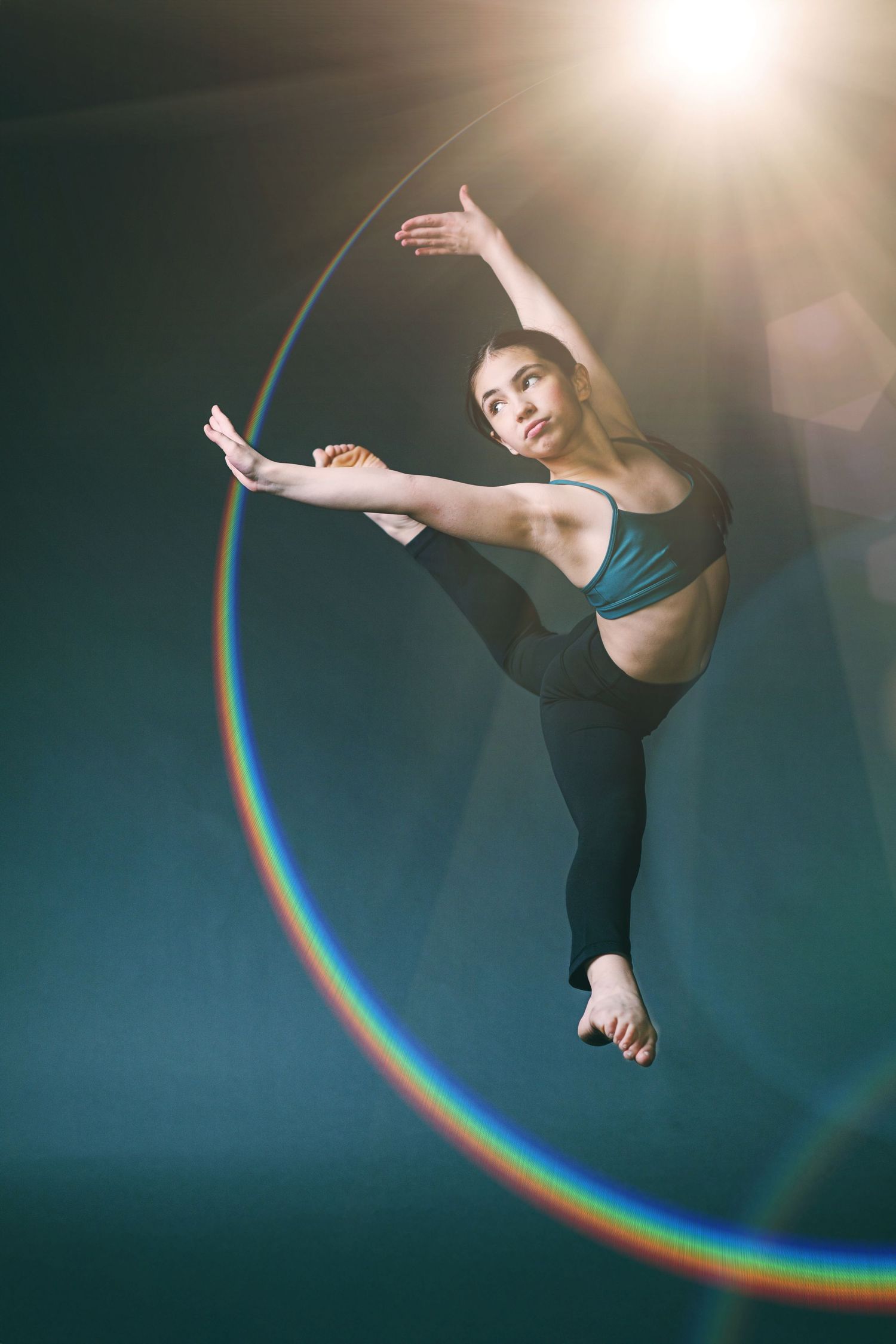 A dancer performs a graceful leap pose with a rainbow arc effect in a dark studio setting.