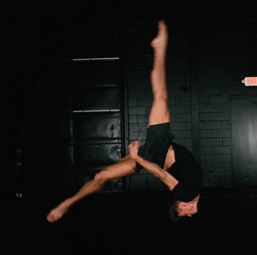 Aerial performer executing an inverted split pose on suspended equipment in a dark studio setting.
