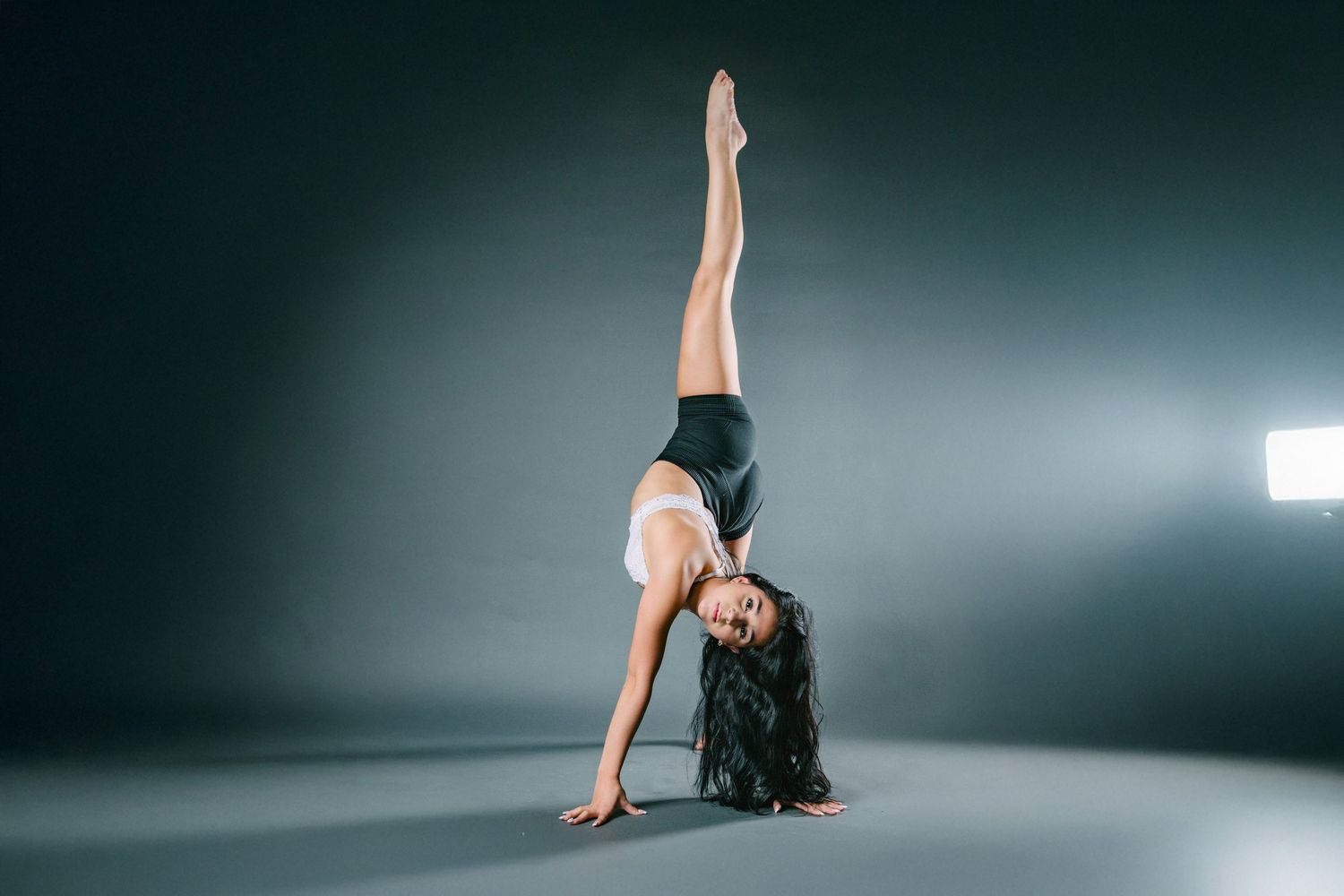 A dancer in a black dress performs a graceful sequence of movements on a dark studio floor with dramatic lighting.