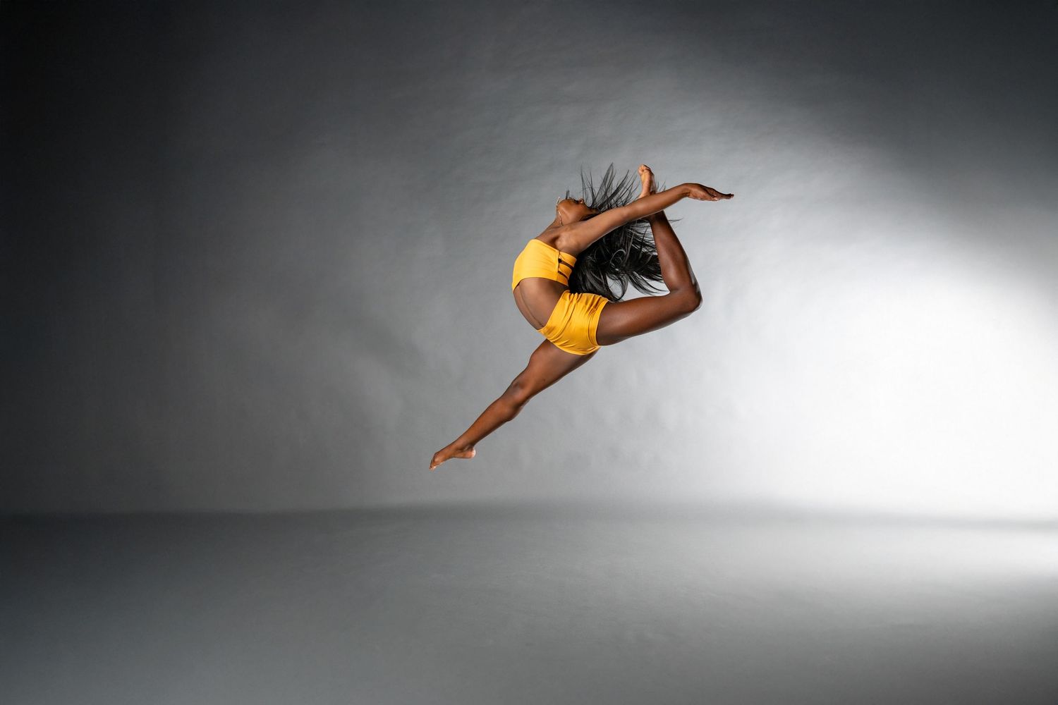Athletic dancer in yellow leotard performs graceful leap against dramatic gray studio backdrop.