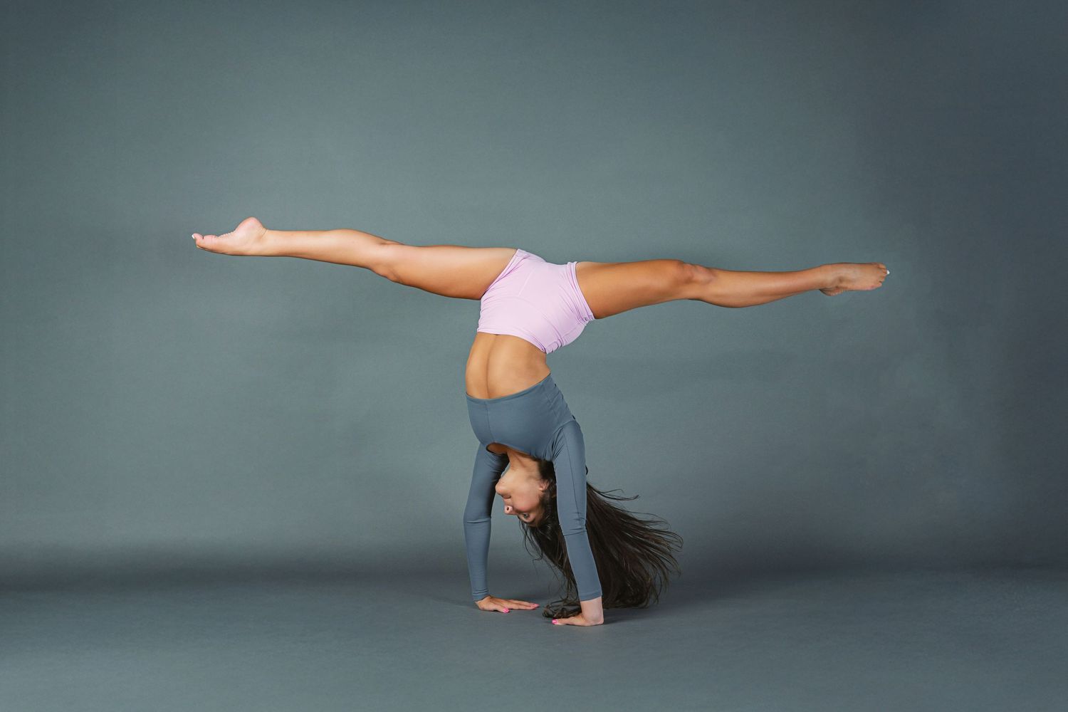 A gymnast performs a graceful handstand split pose against a dark gray studio background.