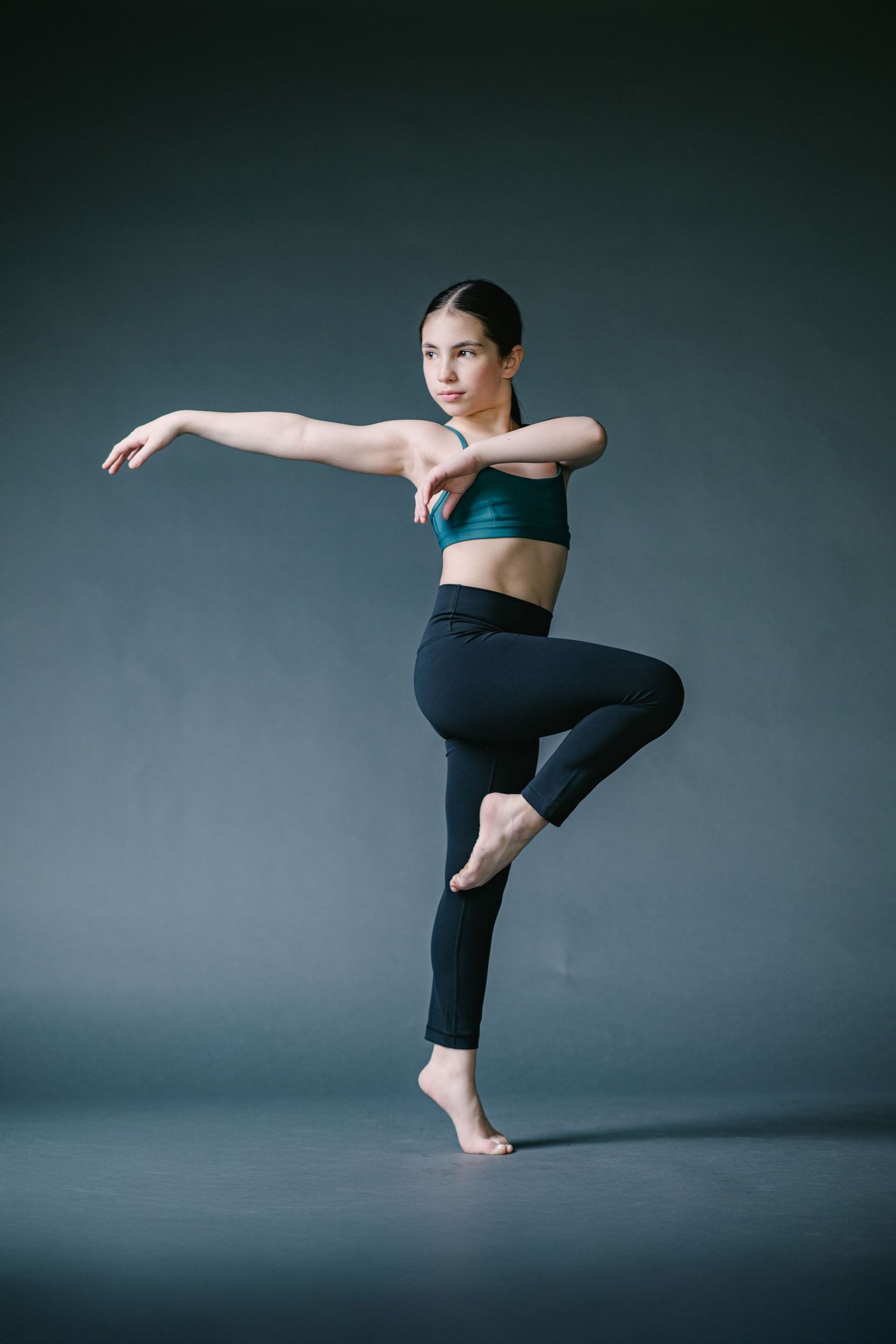 Contemporary dancer in black athletic wear performs a dynamic balance pose against a dark studio backdrop.