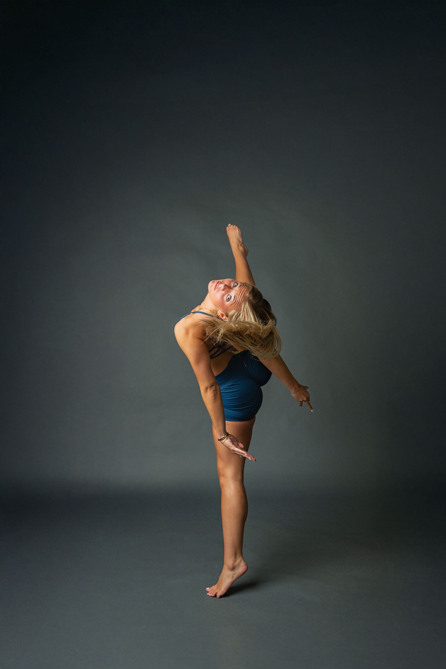 Dancer in blue leotard performs graceful standing split pose against dark studio backdrop.