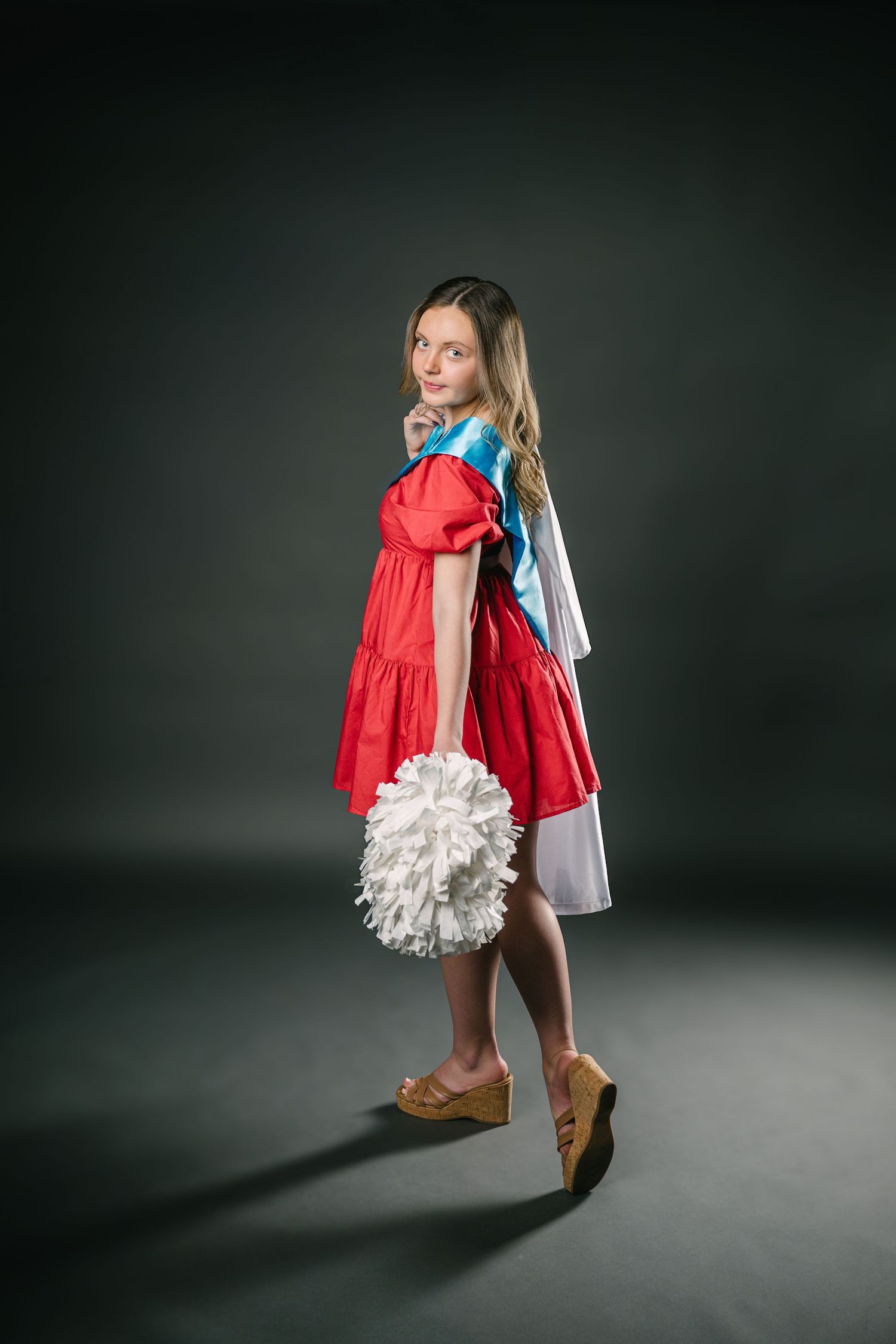 A person in a red cape and white tutu dress twirls in a dramatic studio setting against a dark background.