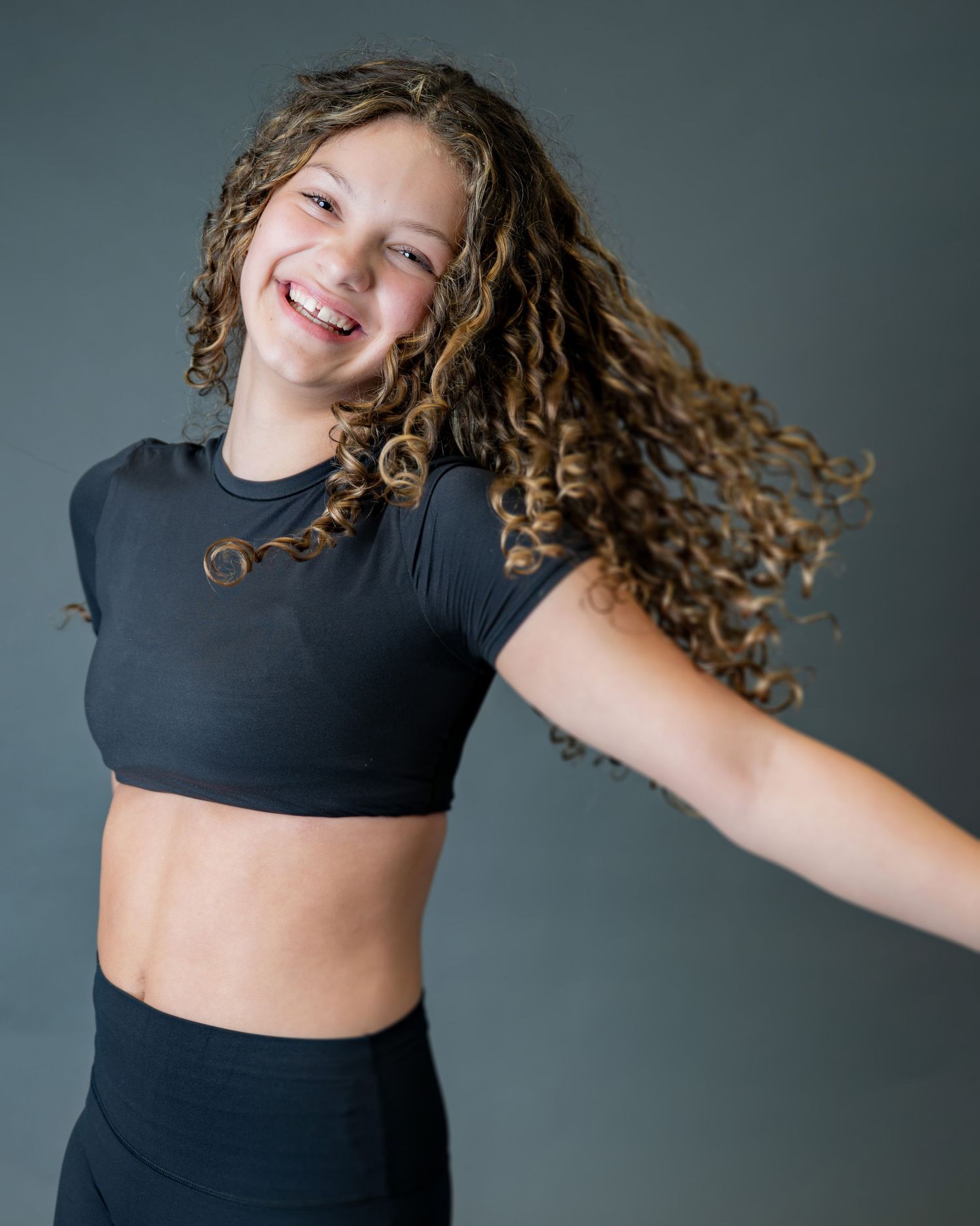 Energetic dancer with curly brown hair wearing black crop top and leggings poses against gray background.