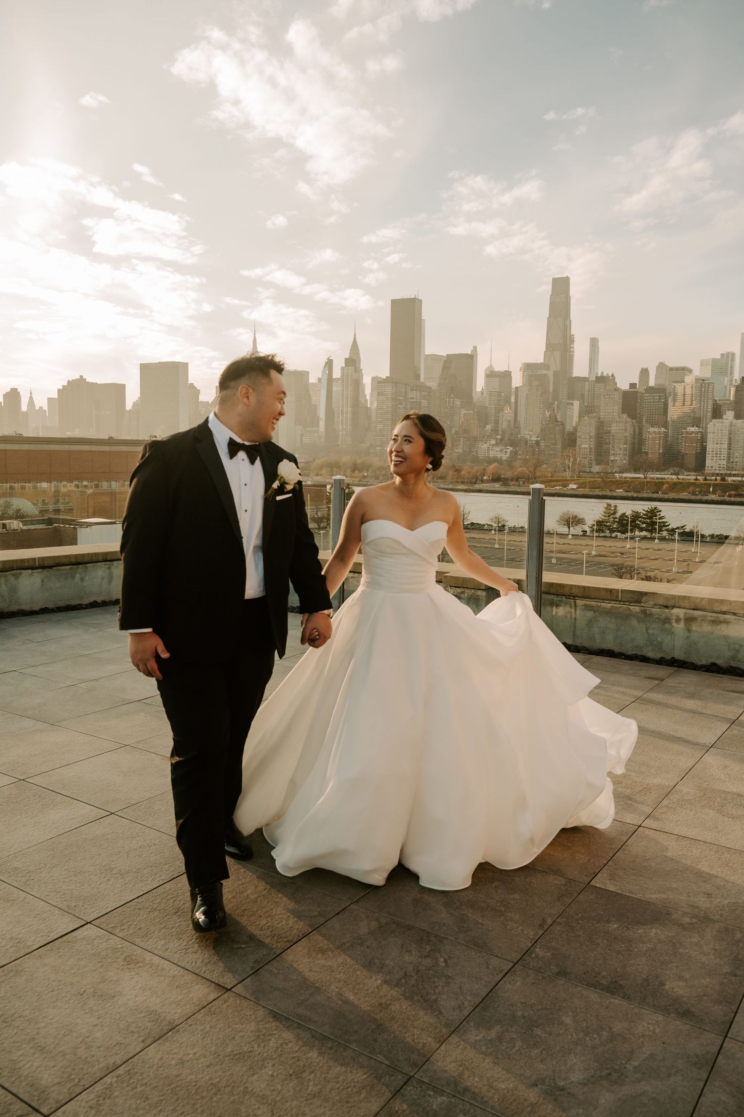 A couple in formal wedding attire walks on a rooftop terrace with the New York City skyline visible in the background.