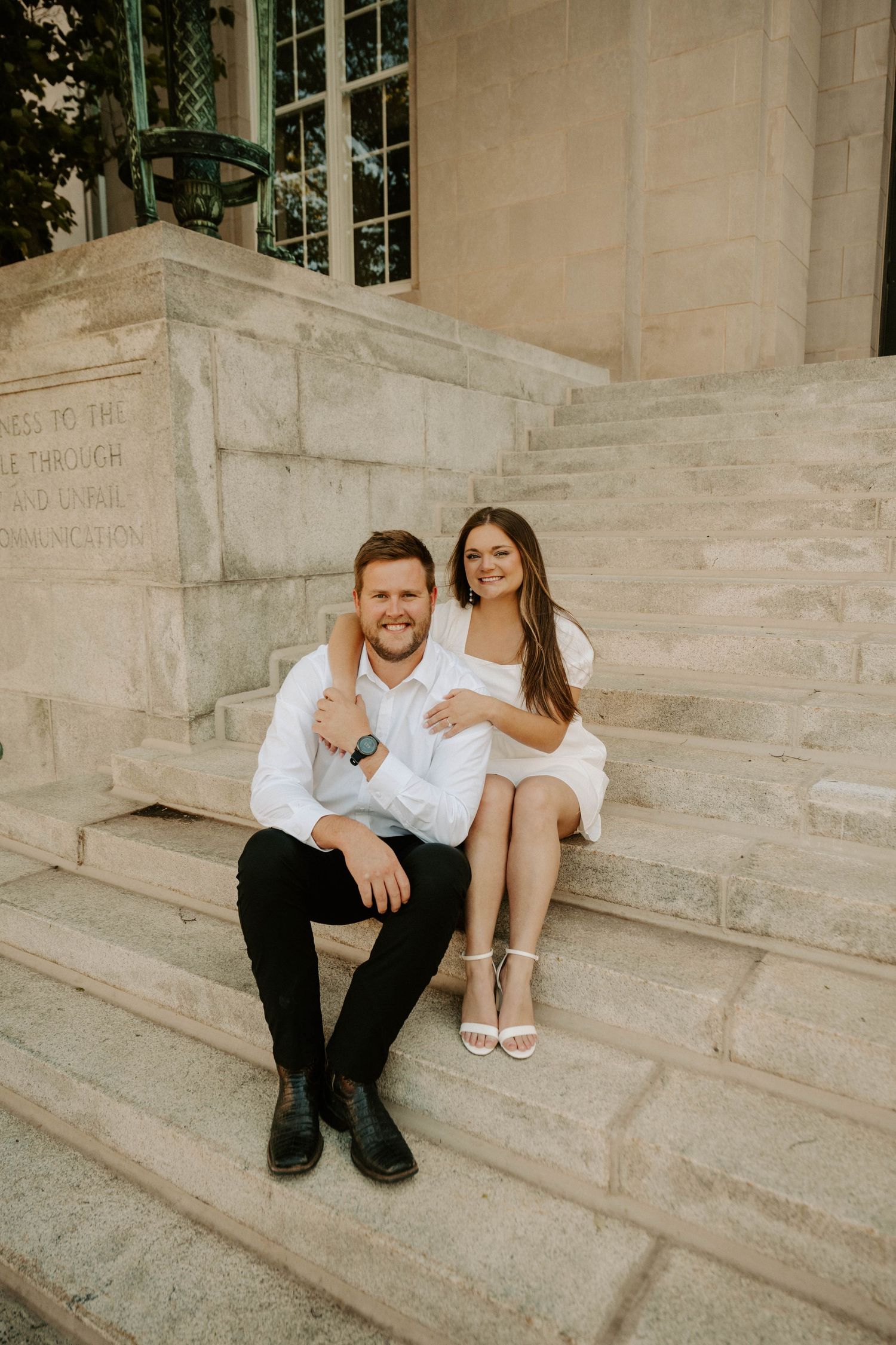 A couple sits together on concrete steps outside a building wearing white shirts and smiling in a romantic engagement photo.