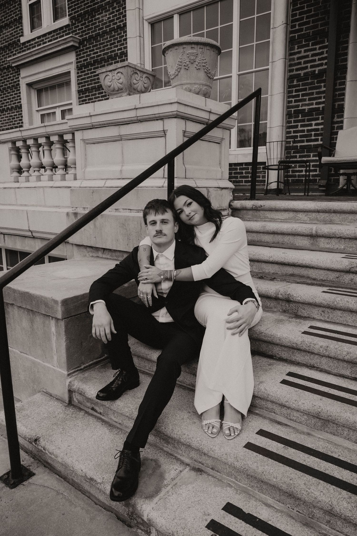 A couple lounges casually on stone steps outside a brick building in a romantic black and white photo series.