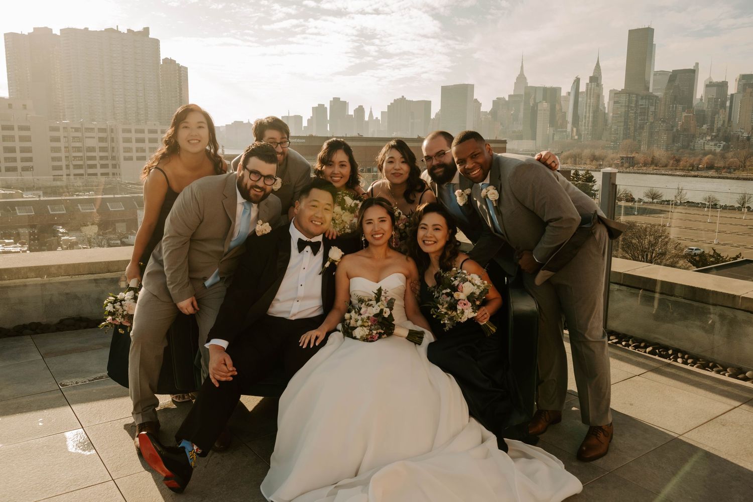 Wedding party poses together on urban rooftop with New York City skyline in background during sunset.
