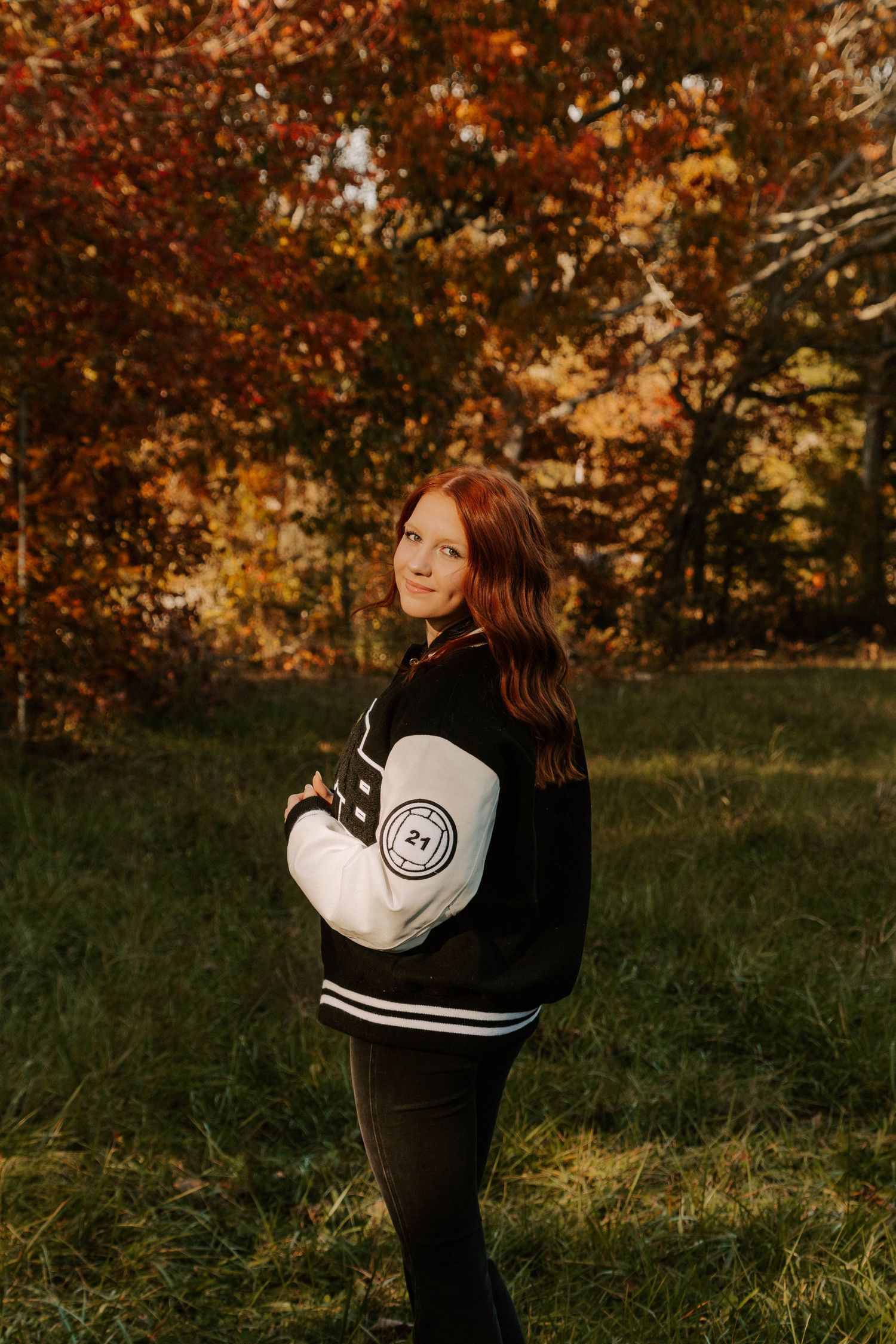 A person wearing a black and white varsity jacket stands in a park with autumn trees displaying vibrant fall colors.