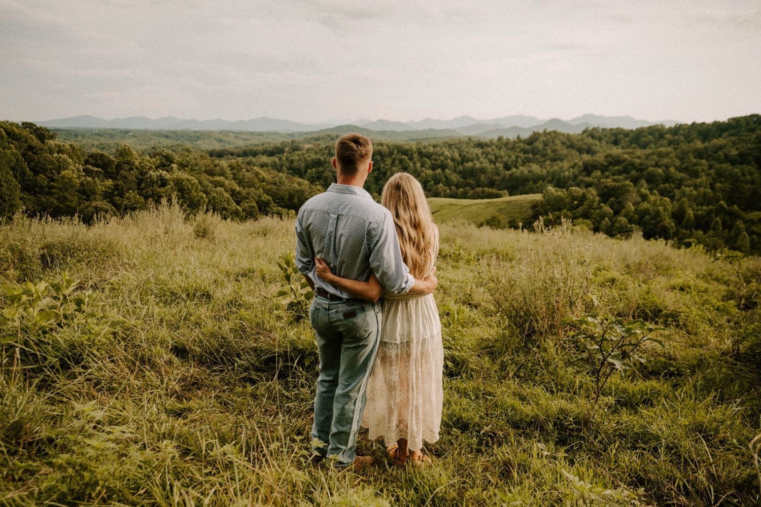 Couple stands in a lush green meadow overlooking rolling hills on a cloudy summer day.