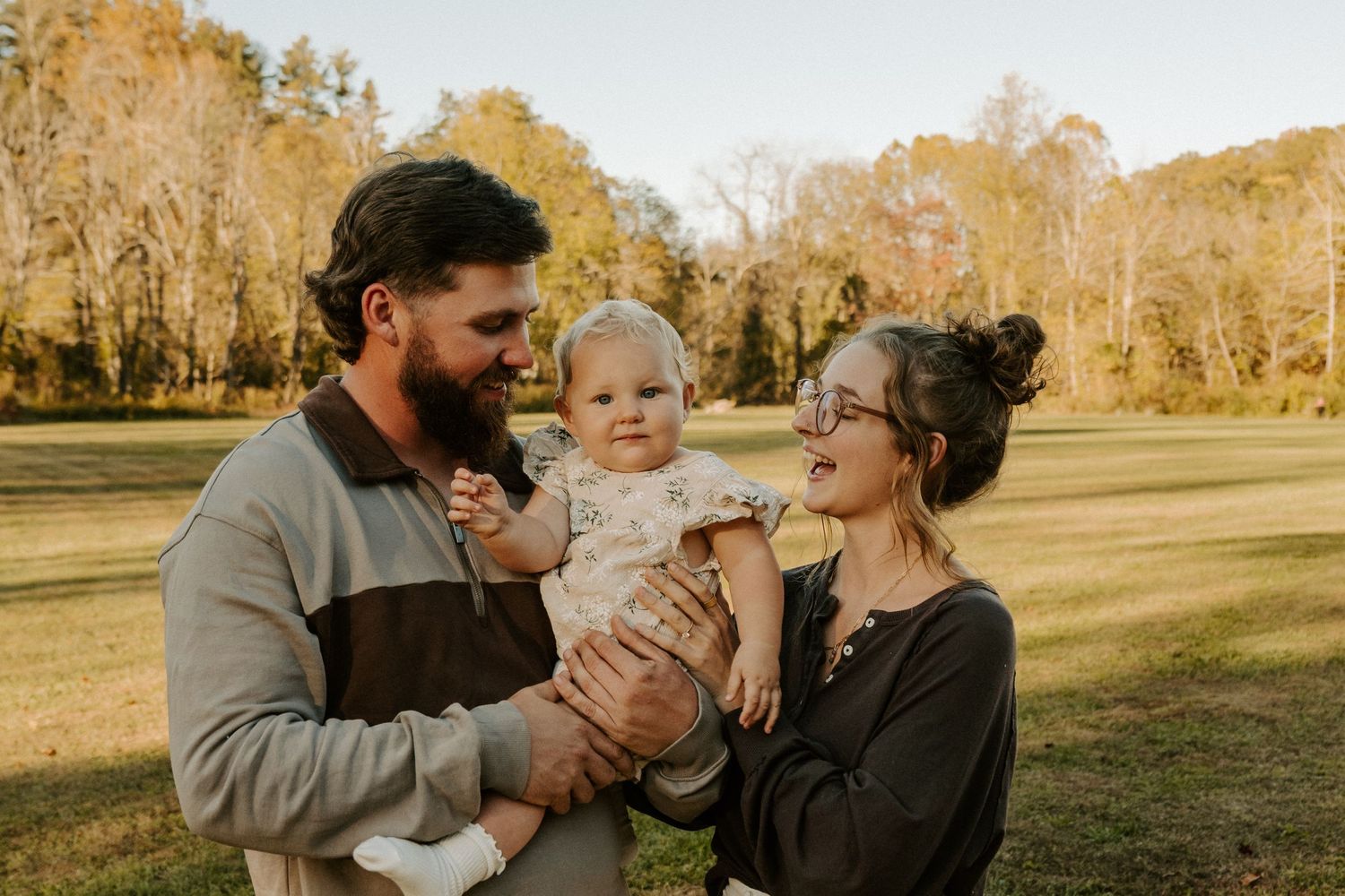 A family shares a tender moment together in a sunny autumn field with golden trees in the background.
