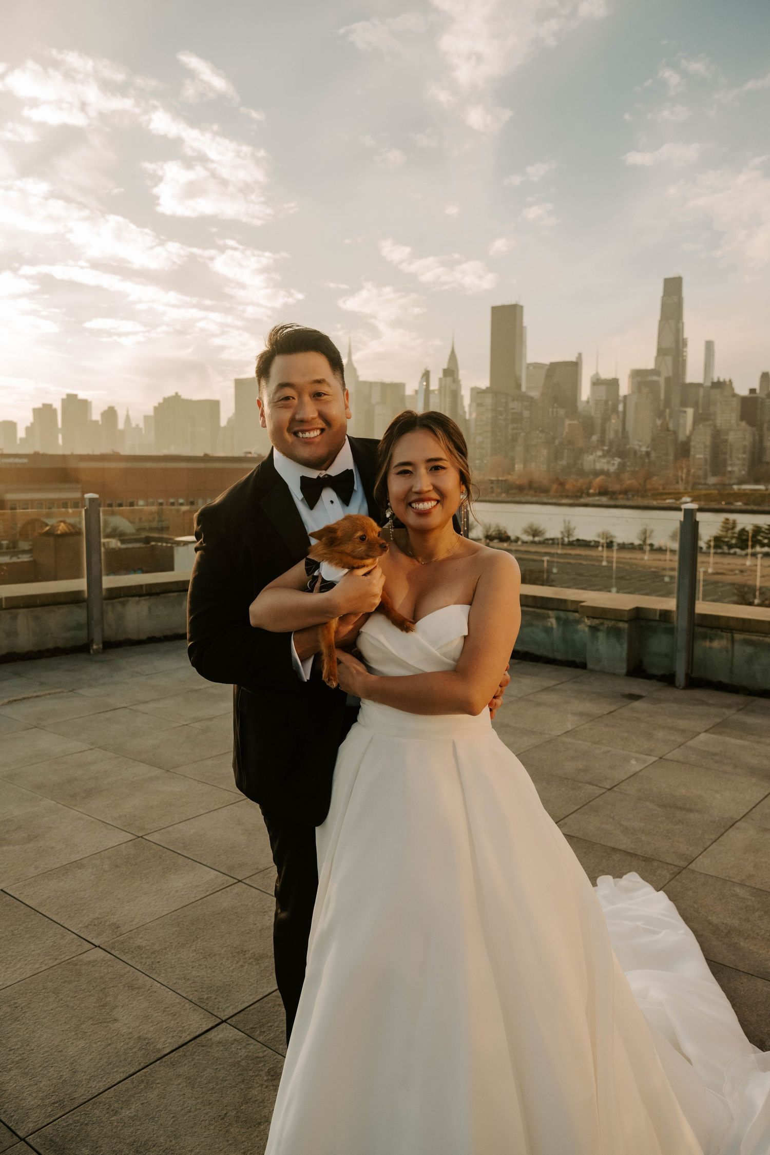 A couple in wedding attire shares a playful moment on a rooftop with the New York City skyline in the background.