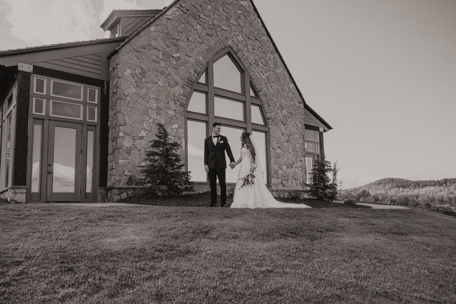 Elegant stone church wedding venue with two people standing on a grassy hill in black and white.