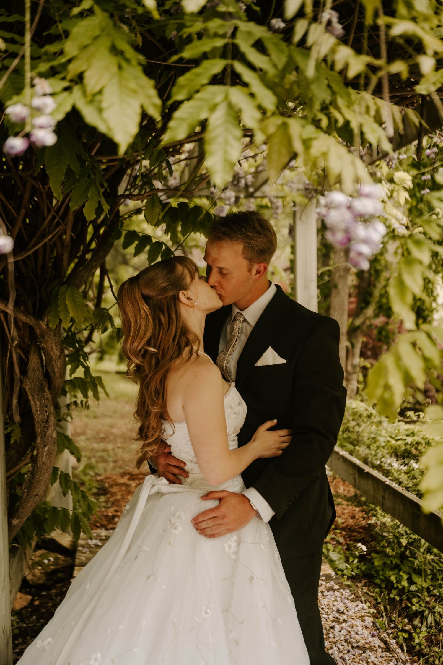 A romantic wedding photo with a strapless white gown and dark suit embracing under a garden arbor with wisteria blooms.