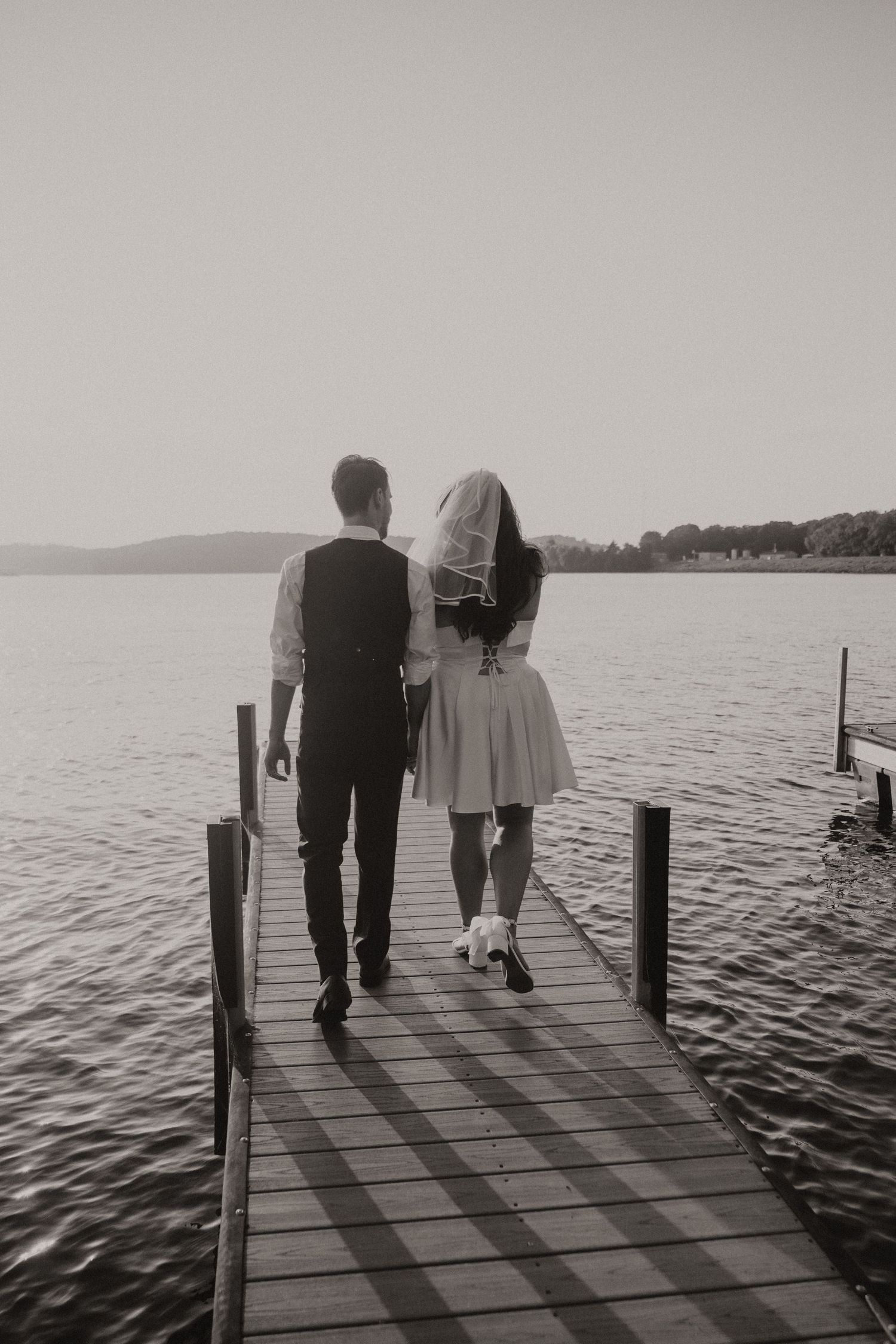 A couple walks together on a wooden dock over a serene lake at sunset in black and white photography.