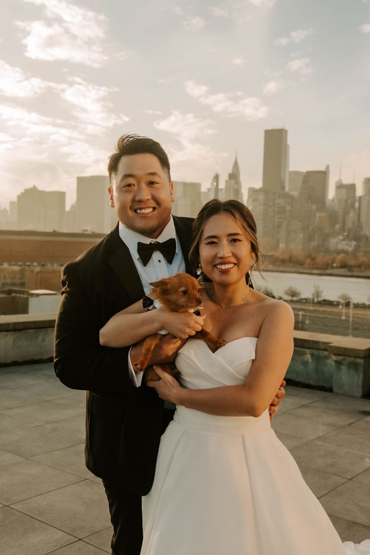 A joyful wedding couple poses with their small dog on a rooftop with the NYC skyline in the background.