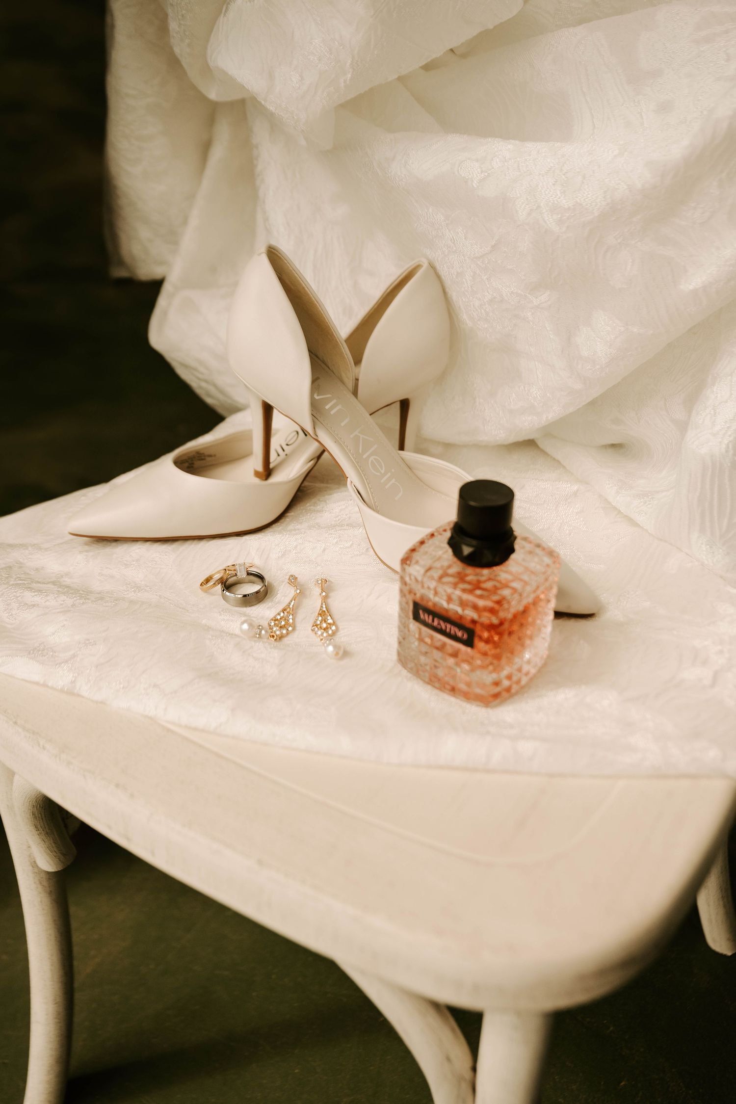 Elegant bridal accessories including white stiletto heels, perfume bottle and jewelry arranged on white table.