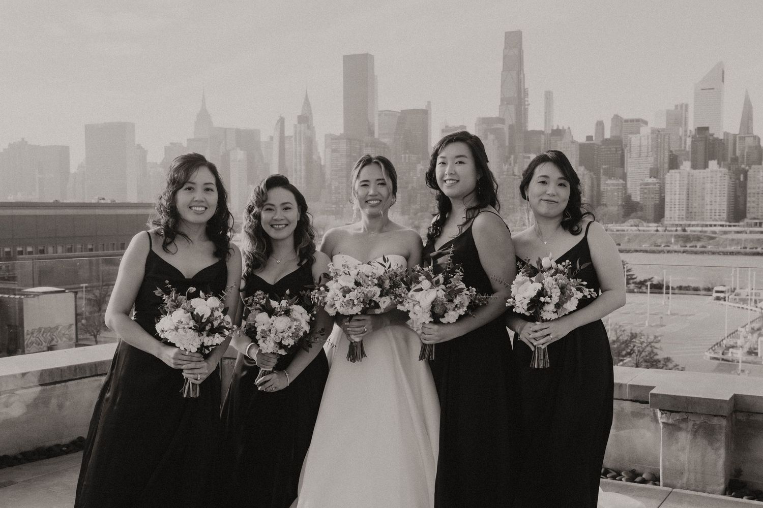 Bridesmaids and bride pose together on urban rooftop with city skyline in background during wedding photoshoot.