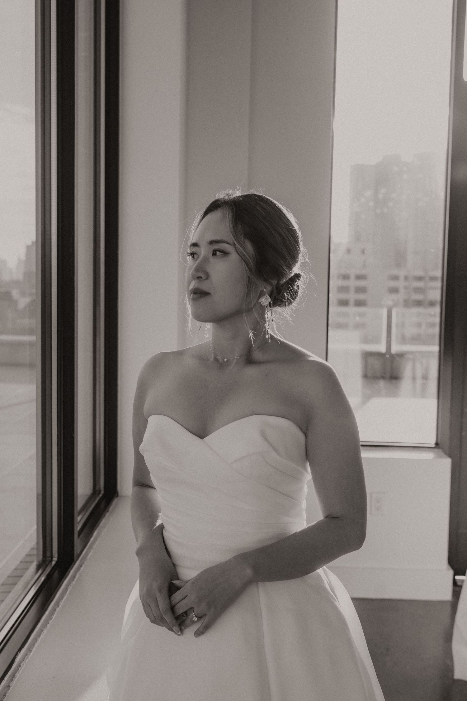 Black and white portrait of a bride in a strapless white wedding dress gazing out a window in soft natural light.