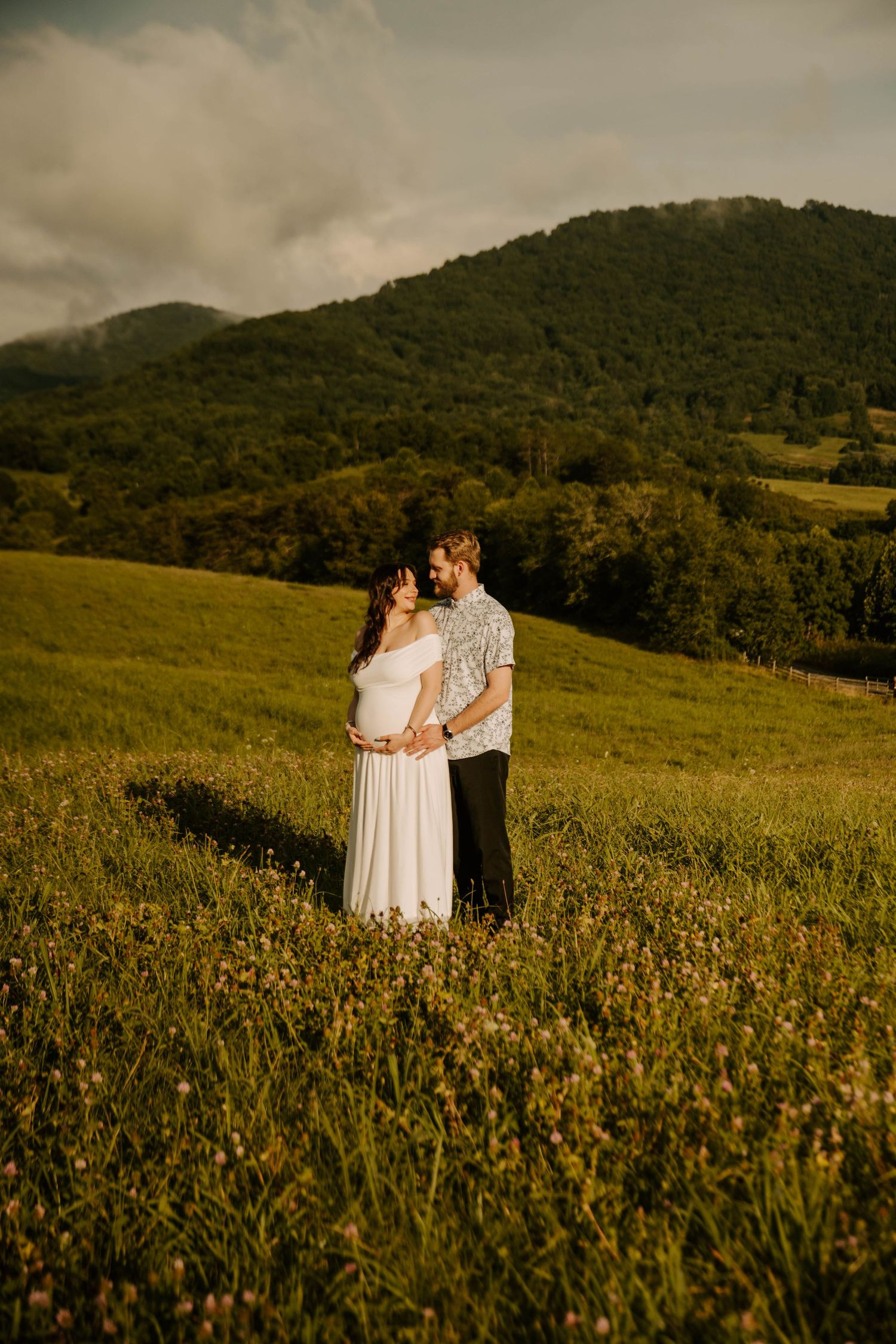 Two figures embrace in a golden wildflower meadow at sunset with rolling green hills in the background.