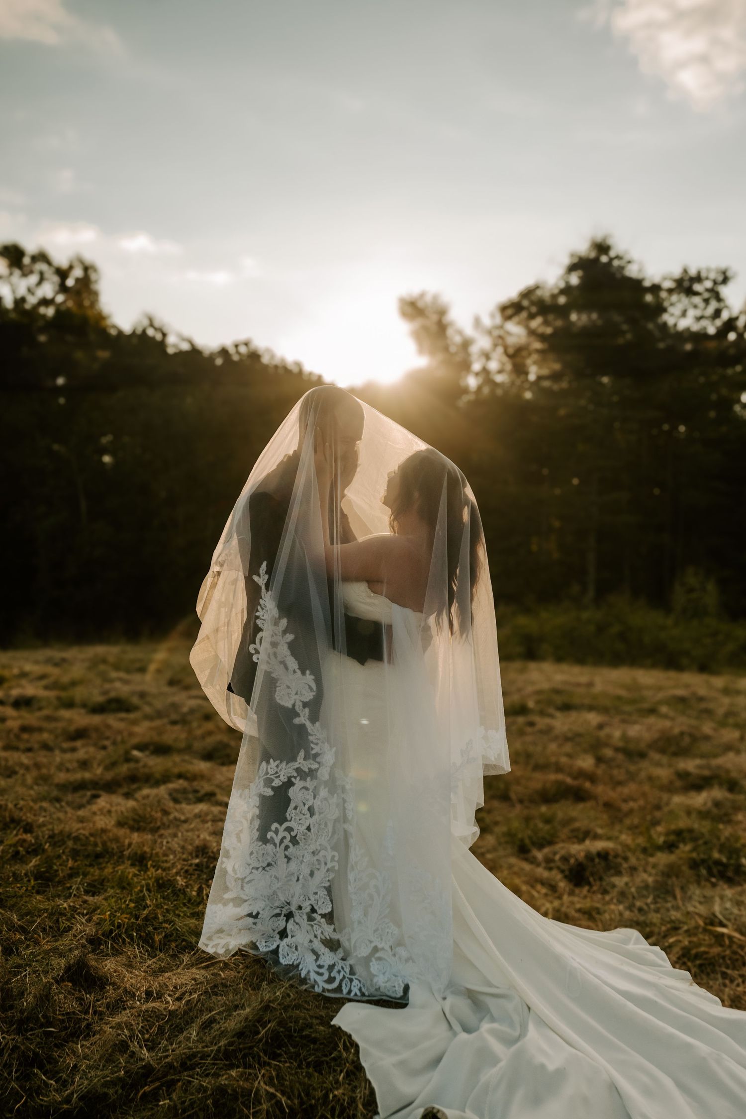 A flowing white wedding veil and dress billows in the wind against a golden sunset in a rustic field.