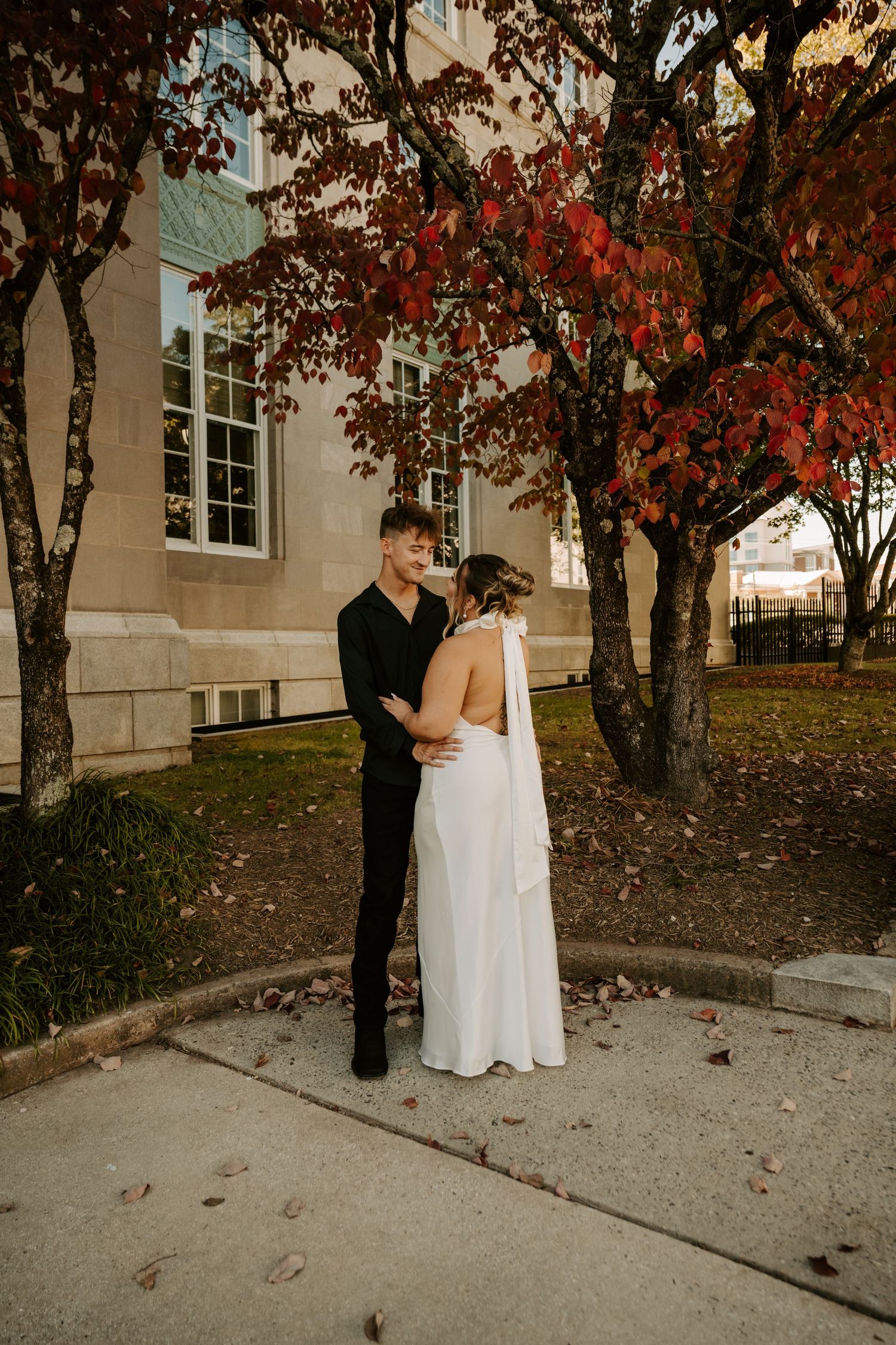 Two people share a tender moment under autumn trees with red leaves on a walkway next to a white building.