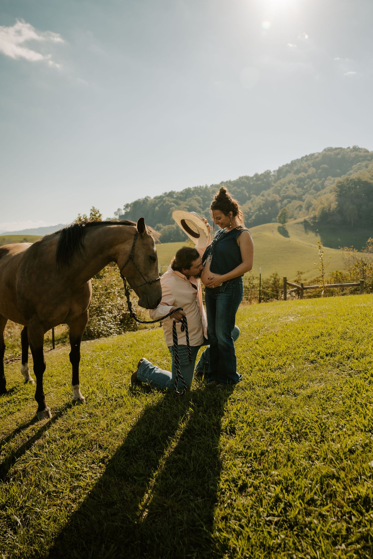 A horse stands in a sunlit mountain pasture with two people during golden hour, casting long shadows on the grass.