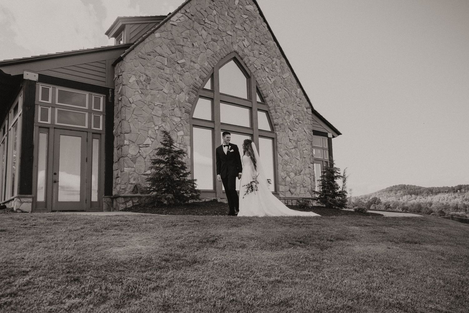 A modern stone house with large windows sits on a grassy hillside overlooking a scenic mountain landscape.