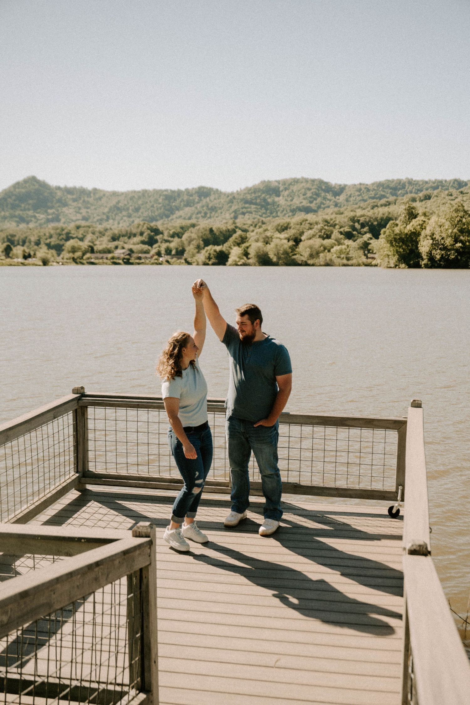 Couple dancing on a wooden dock overlooking a serene lake surrounded by rolling hills on a sunny autumn day.