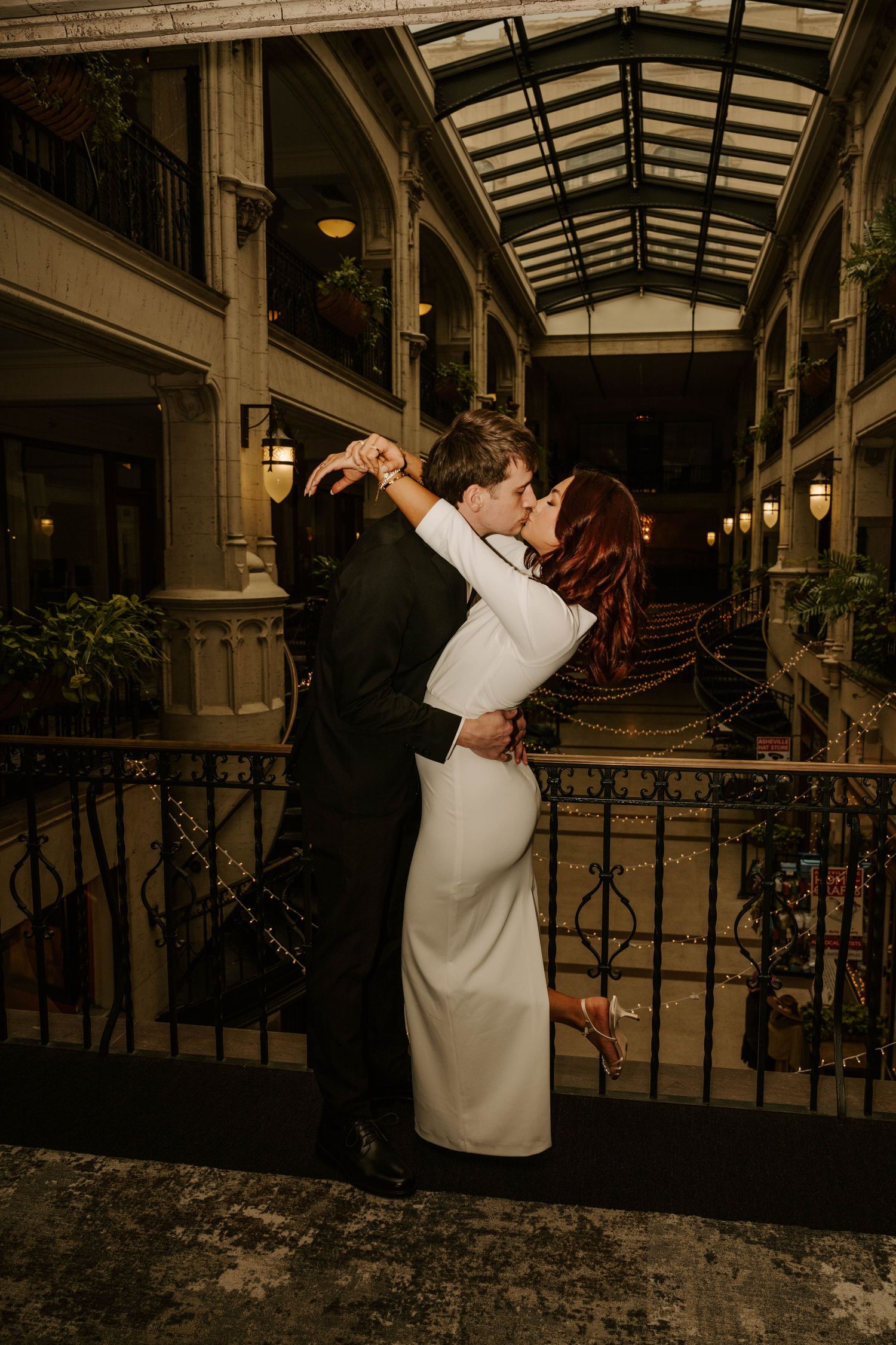 A romantic wedding couple embraces in a covered walkway with elegant architecture and warm lighting at dusk.