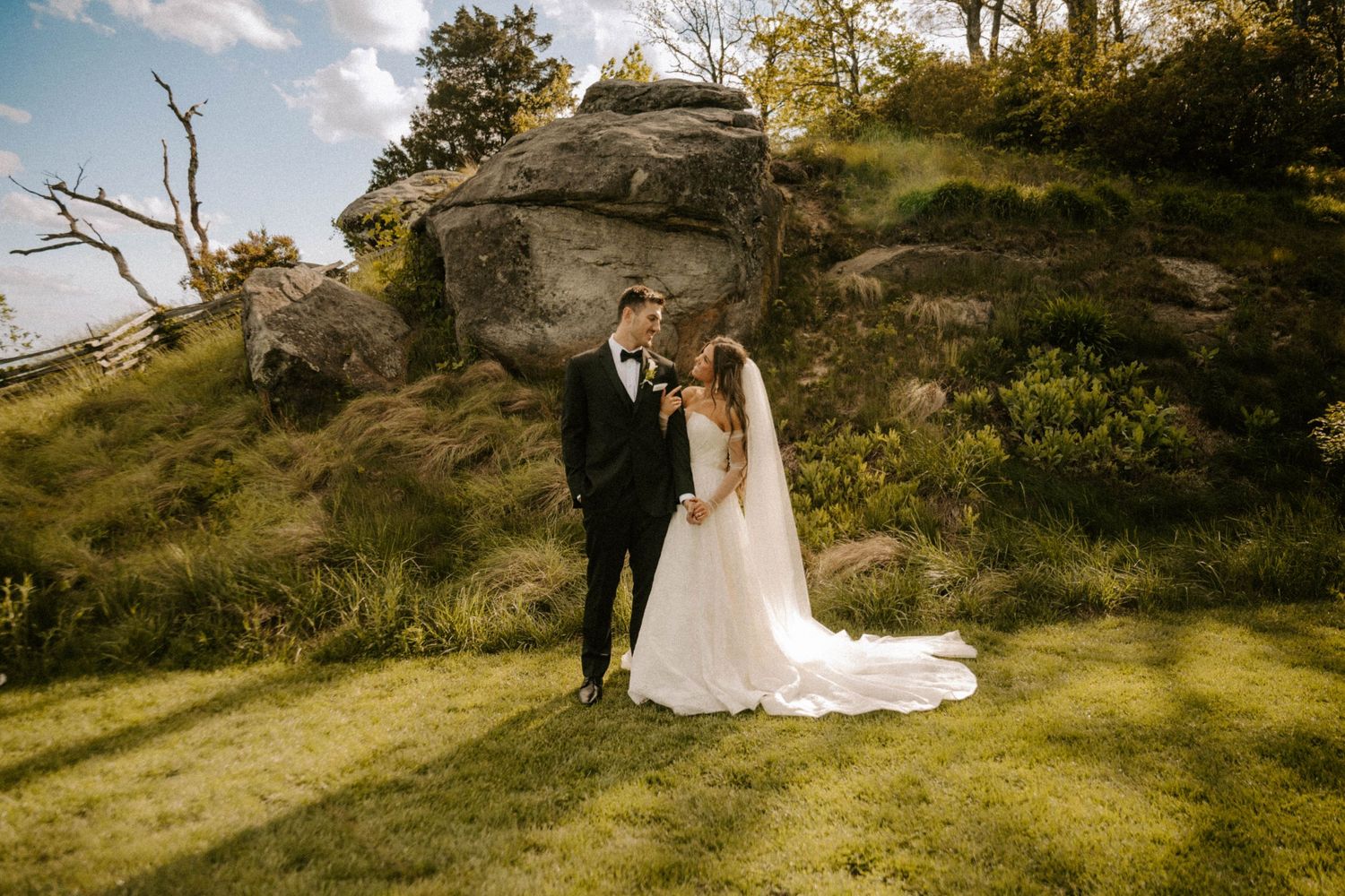 Two people in wedding attire pose on a rocky hilltop with green grass and natural scenery in the background.